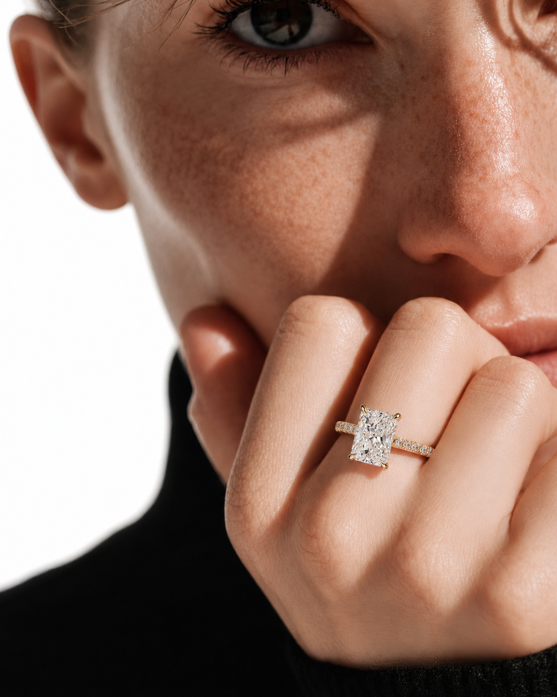 color_10K_YG, color_14K_YG &&
A close-up lifestyle shot of a model wearing a yellow gold engagement ring with a rectangular baguette-cut diamond. The ring is set on the model's ring finger, with dramatic side lighting casting shadows across her face and hand.