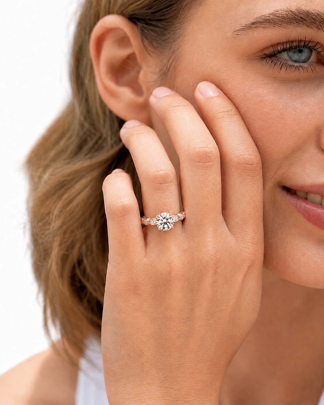 color_10K_RG, color_14K_RG &&
A close-up lifestyle shot of a woman wearing a rose gold diamond ring with a round-cut center stone and smaller side stones on her finger, with her hand resting against her cheek.