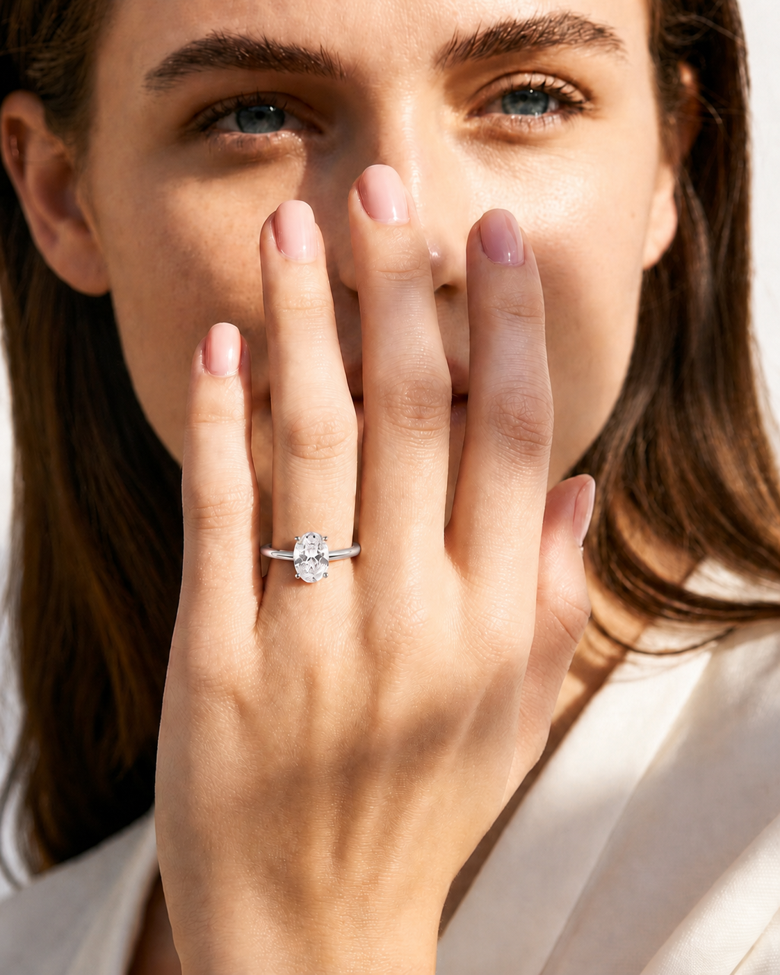 color_925_SIL,color_935_ARG,color_10K_WG, color_14K_WG &&A close-up lifestyle shot of a woman holding her hand to her face, showcasing a silver engagement ring with a round-cut clear gemstone. The ring is centered on her ring finger, with warm, bright lighting and a soft-focus background.