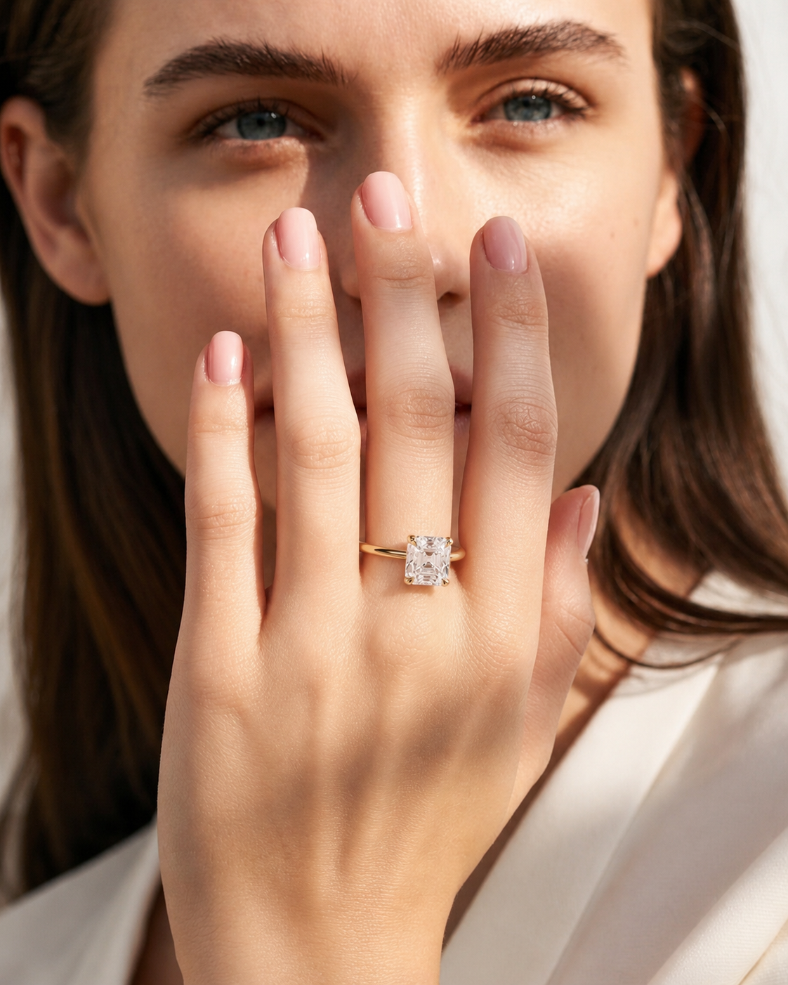color_10K_YG, color_14K_YG &&A close-up lifestyle shot of a woman holding her hand near her face, wearing a gold ring with a rectangular clear gemstone on her ring finger.