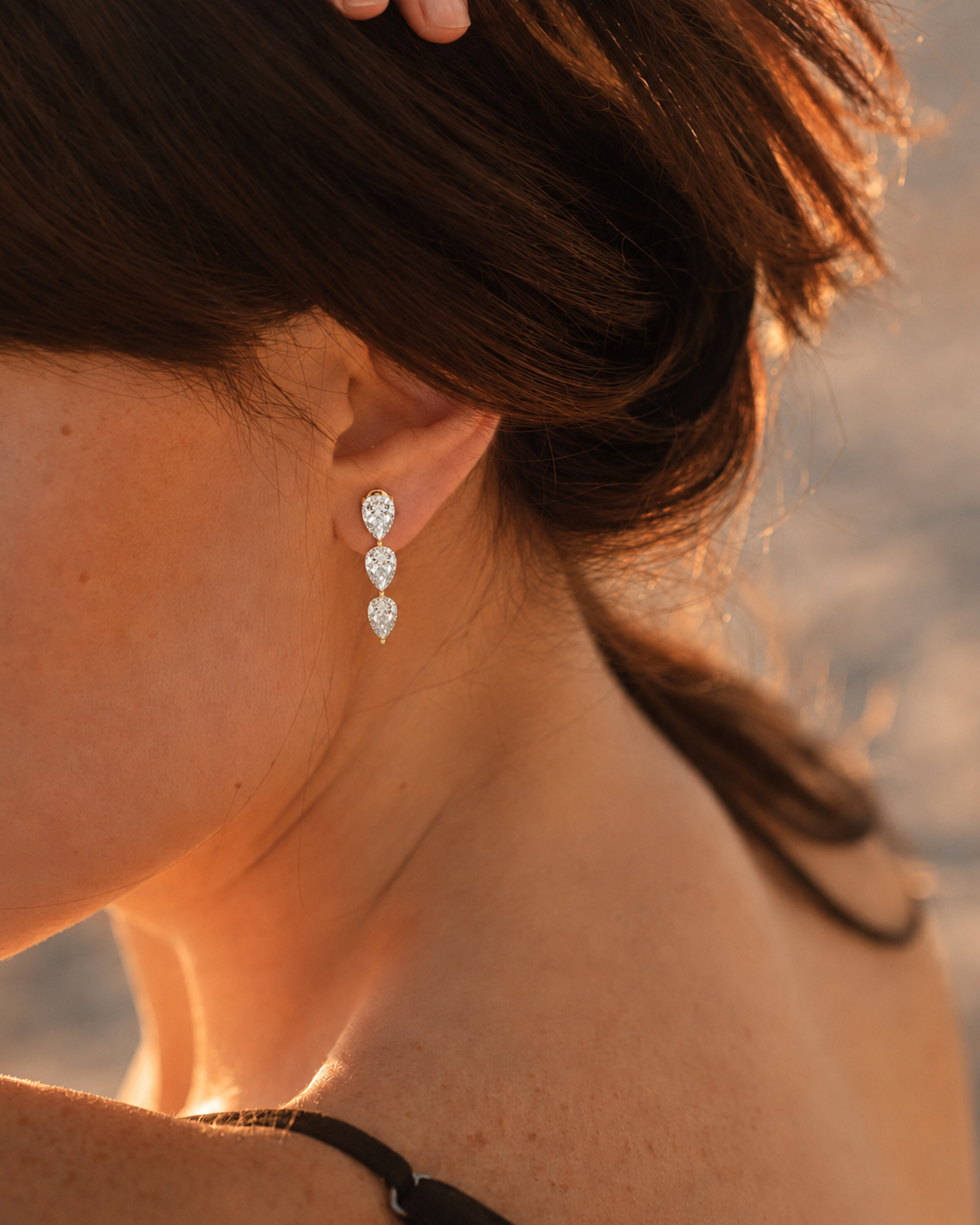 color_10K_YG, color_14K_YG &&A close-up lifestyle shot of a woman wearing a small square stud earring with a silver-toned frame and clear gemstone, set against a warm, sunlit outdoor background.