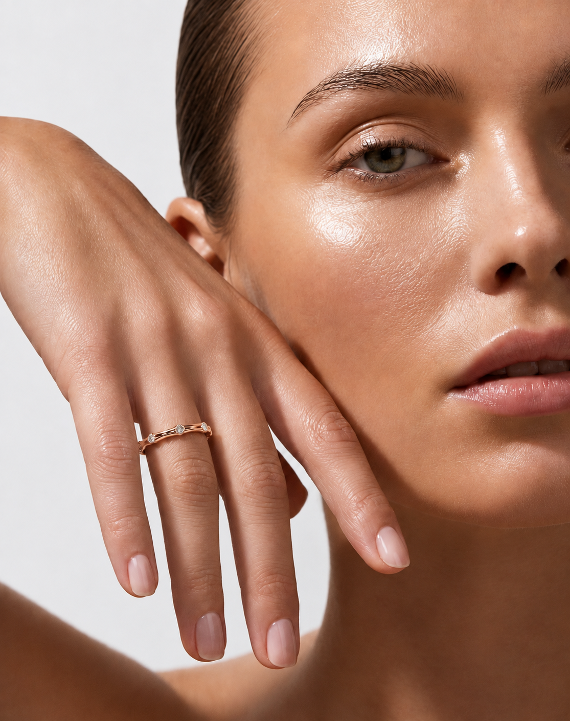 color_10K_RG, color_14K_RG &&	
A close-up lifestyle shot of a model wearing a delicate rose gold band ring with small clear stones on her finger, with her hand resting against her cheek against a light gray background.