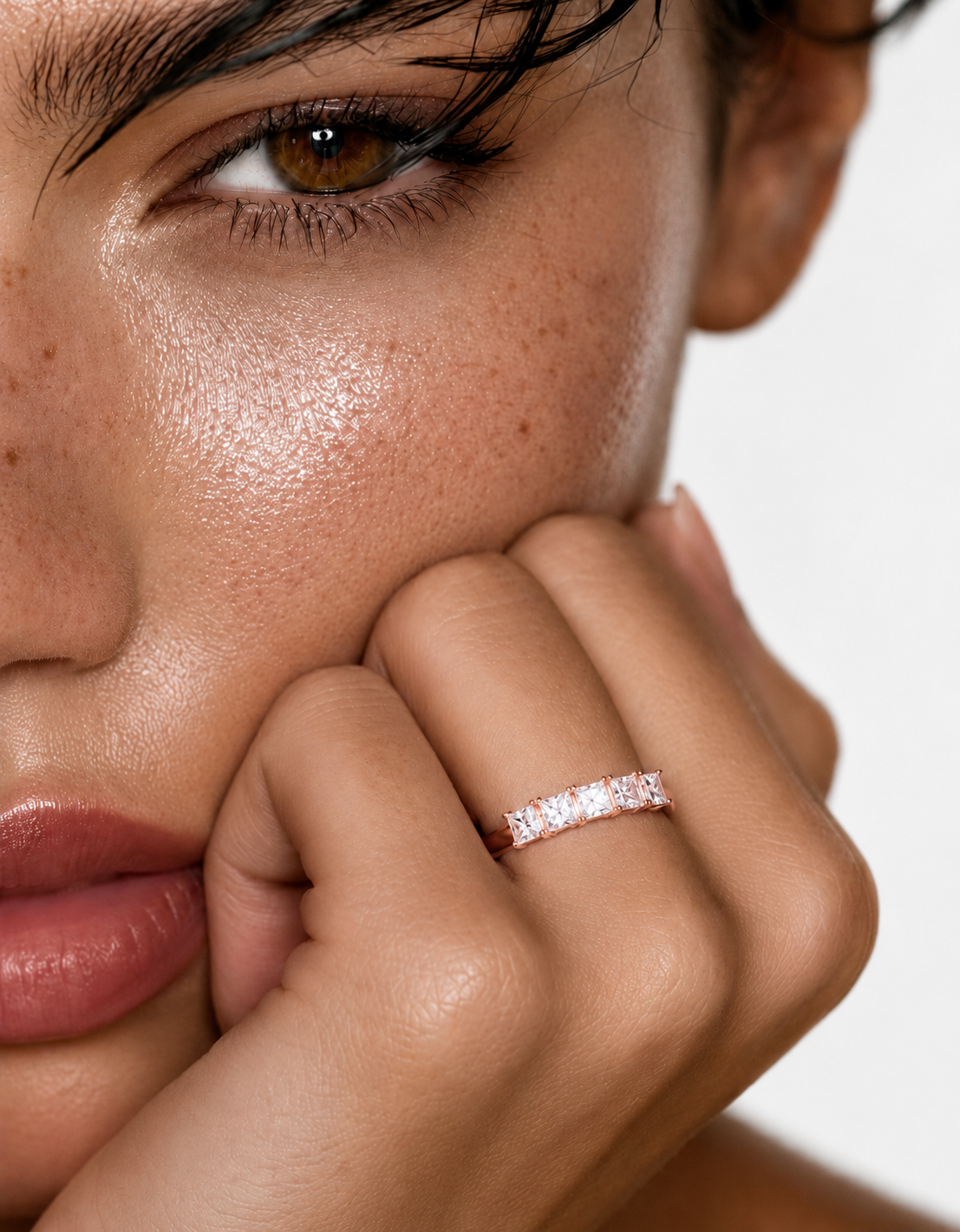 color_10K_RG, color_14K_RG &&	
A close-up lifestyle shot of a model wearing a delicate rose gold eternity ring set with small clear stones on her finger, with her hand resting near her face against a clean white background.
