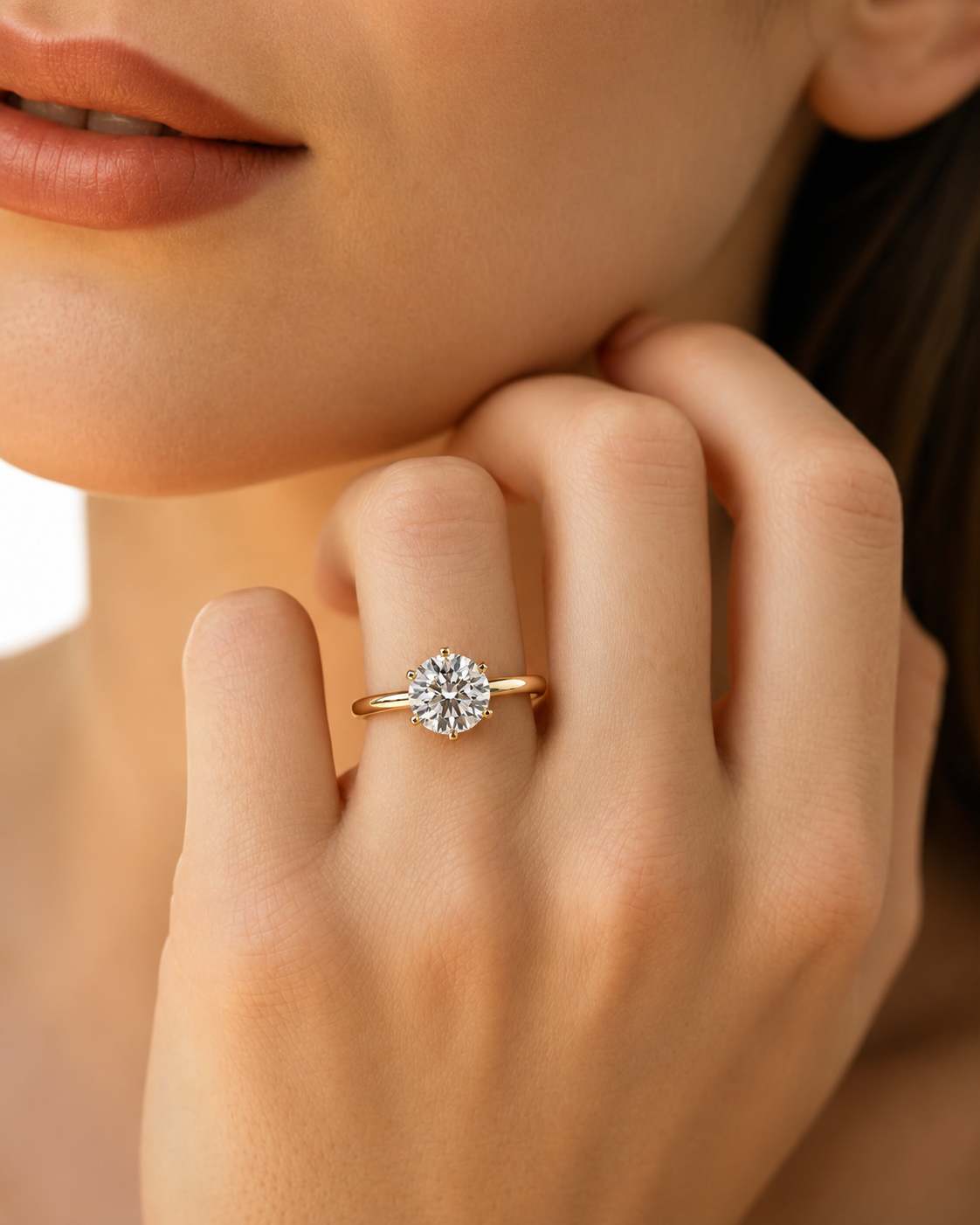 color_10K_YG, color_14K_YG &&A close-up lifestyle shot of a woman's hand wearing a yellow gold ring with a round-cut clear gemstone, set against her cheek and jawline.