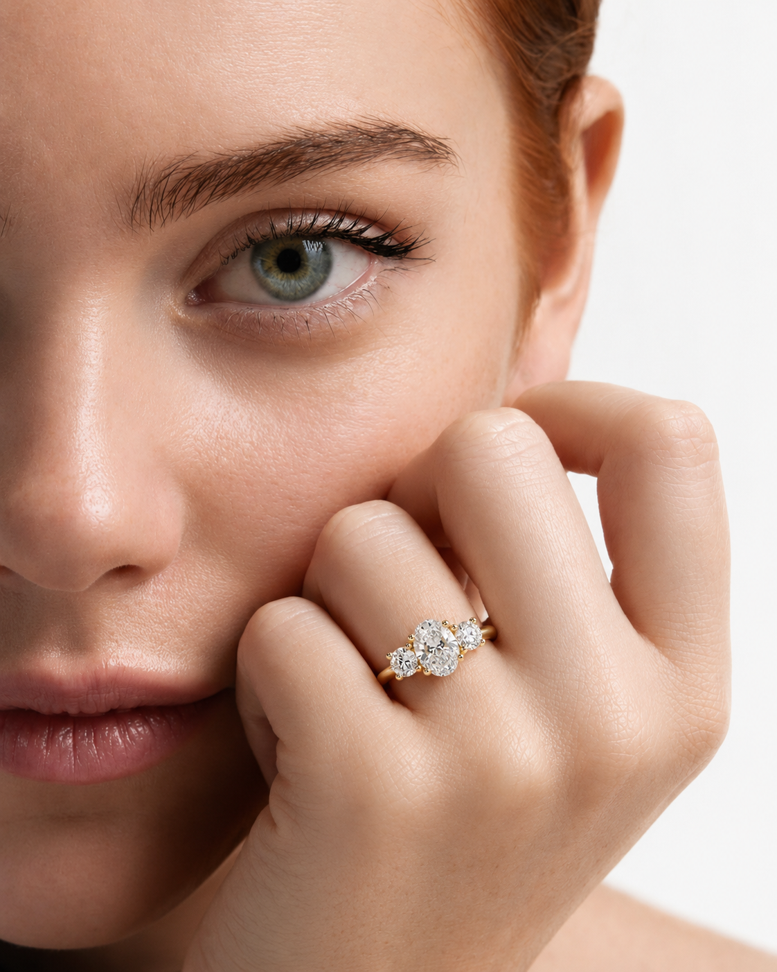 color_10K_YG, color_14K_YG &&A close-up lifestyle shot of a woman with red hair resting her chin on her hand, wearing a yellow gold ring with a three-stone diamond cluster.