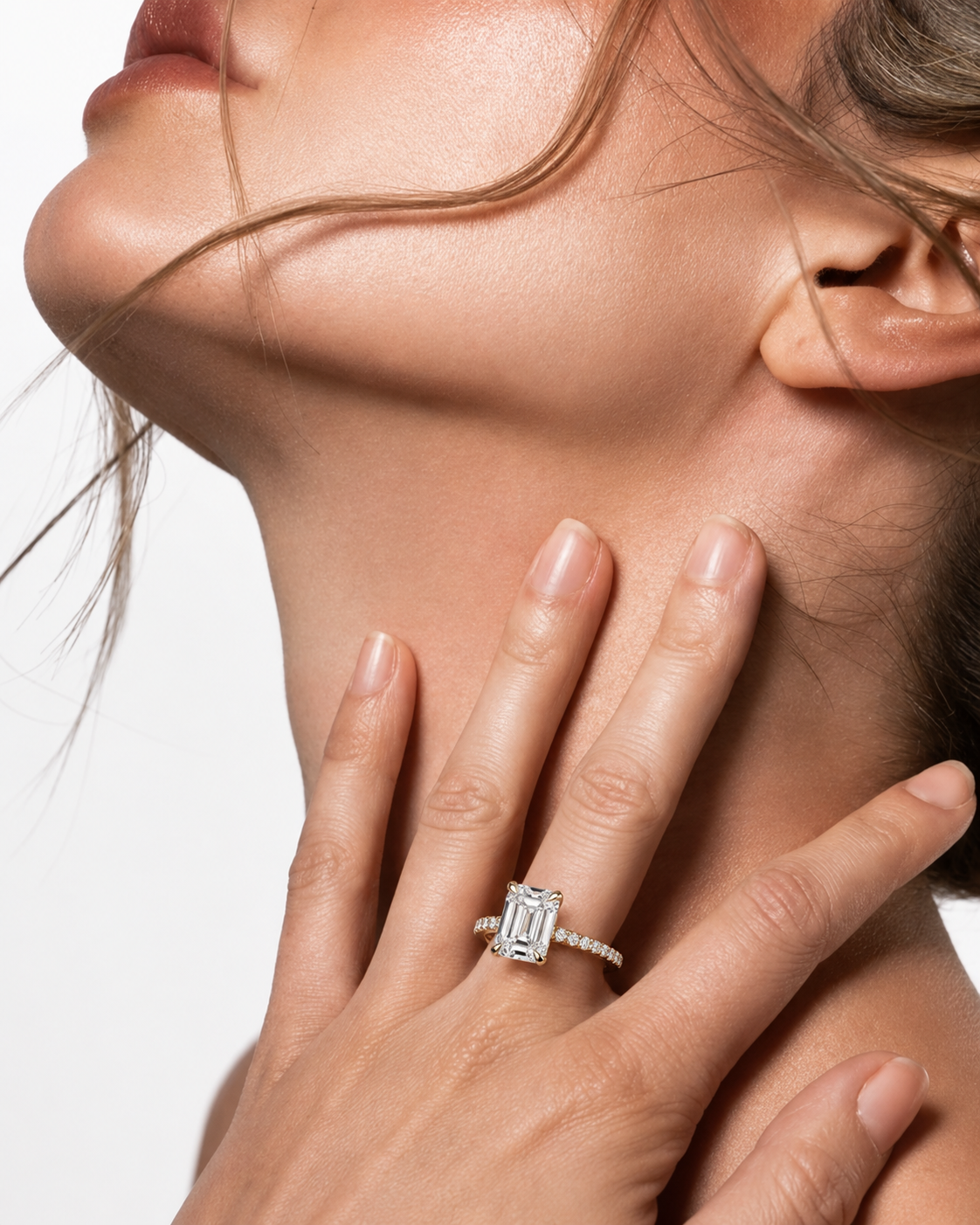 color_10K_YG, color_14K_YG &&A close-up lifestyle shot of a model wearing a silver emerald-cut diamond engagement ring on her finger, with her hand resting on her neck against a clean white background.