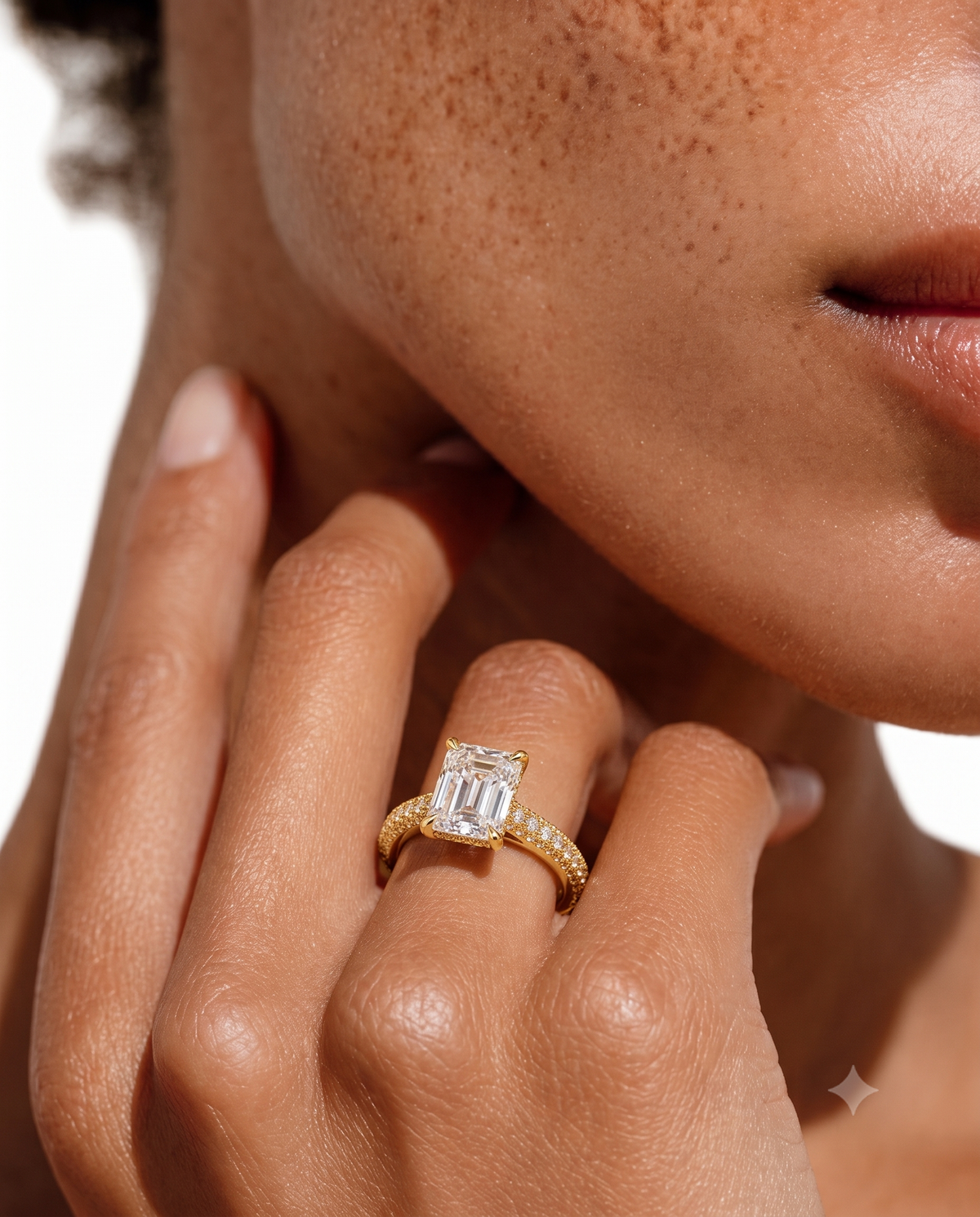color_10K_YG, color_14K_YG &&A close-up lifestyle shot of a model wearing a yellow gold ring with an emerald-cut clear center stone and a pavé band, resting her hand near her face.