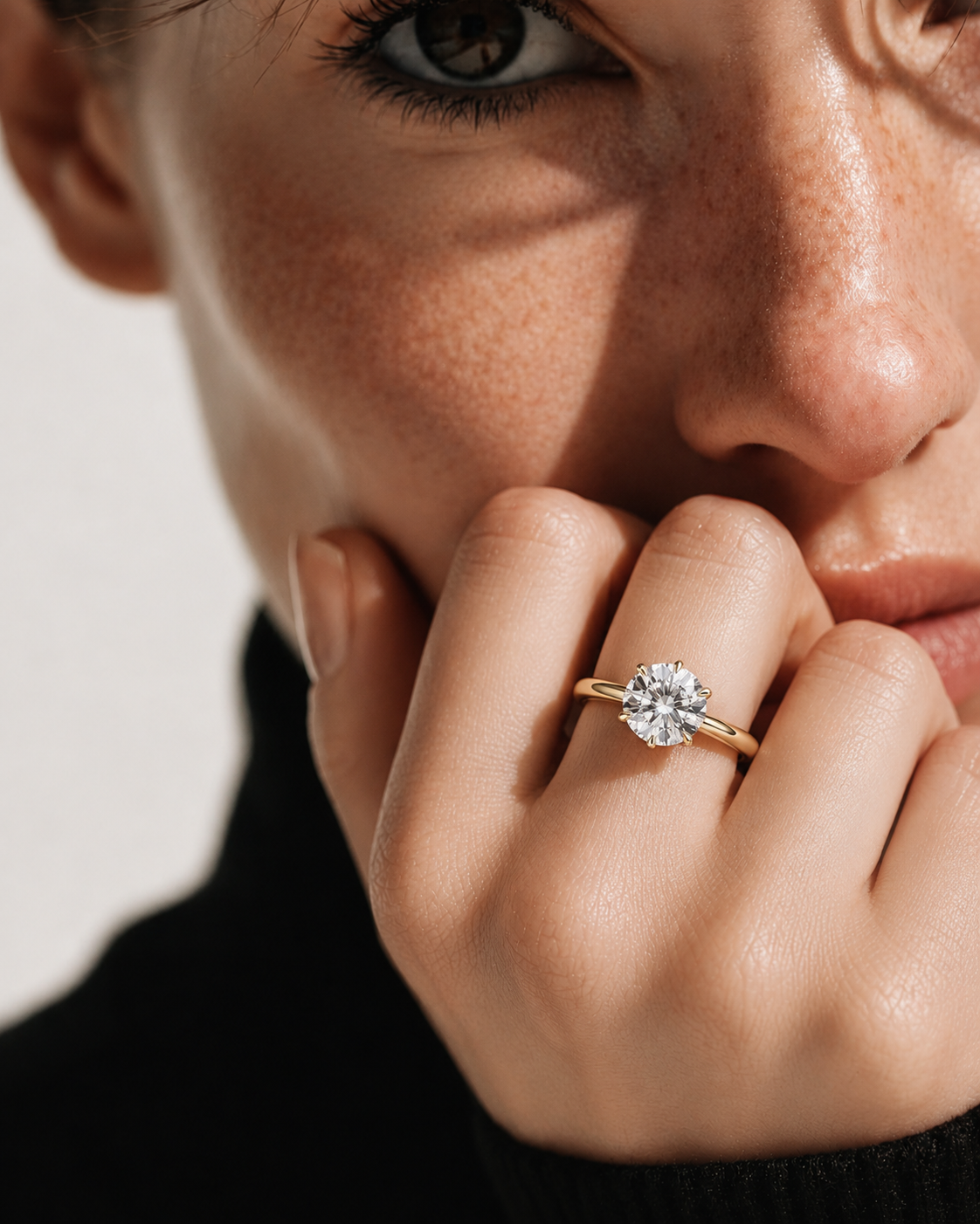 color_10K_YG, color_14K_YG &&A close-up lifestyle shot of a model wearing a yellow gold ring with a round-cut clear gemstone on her finger, resting her chin on her hand against a neutral light background.