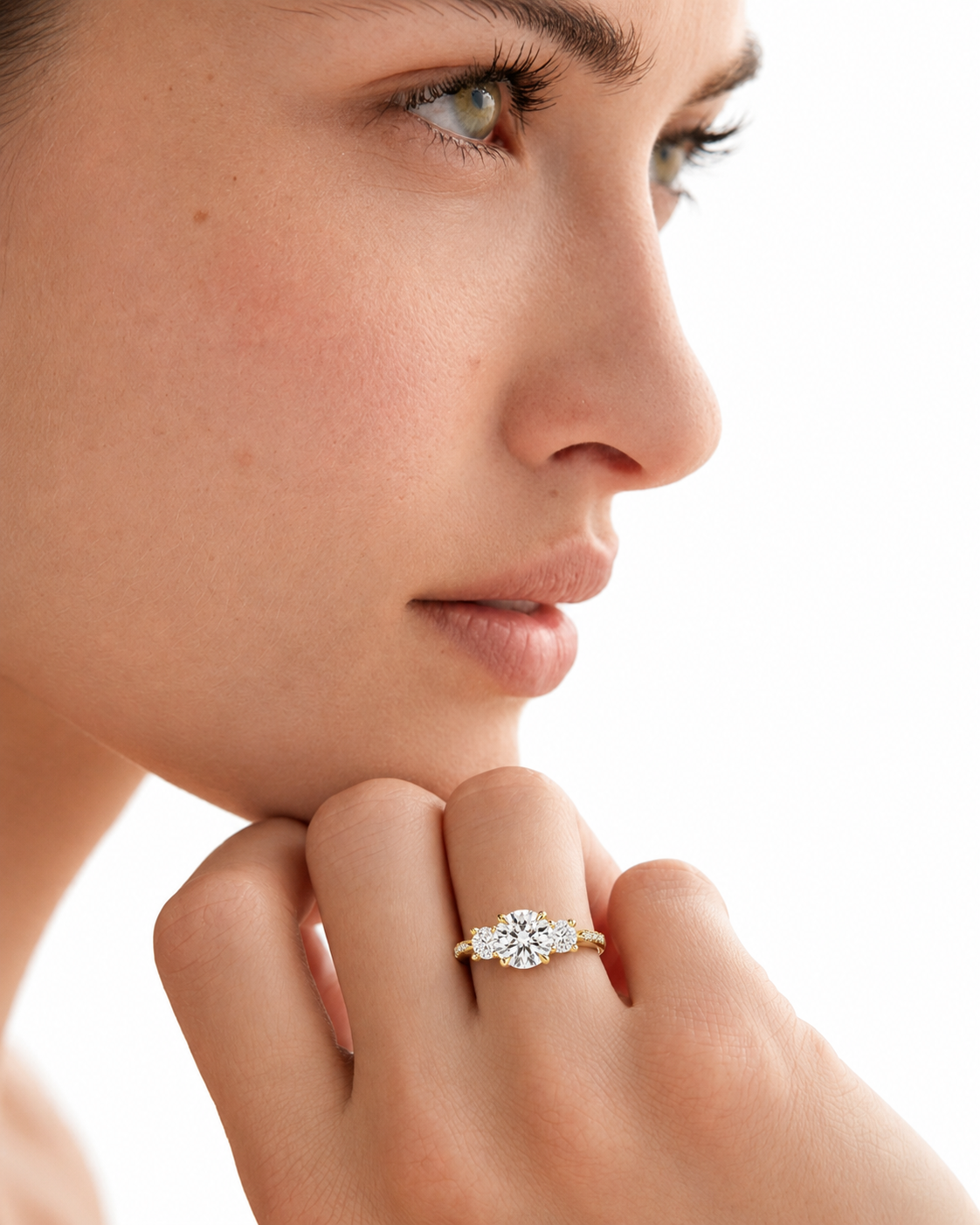 color_10K_YG, color_14K_YG &&A close-up lifestyle shot of a woman's face and hand, wearing a yellow gold ring with a marquise-cut diamond center and smaller round diamonds on either side.