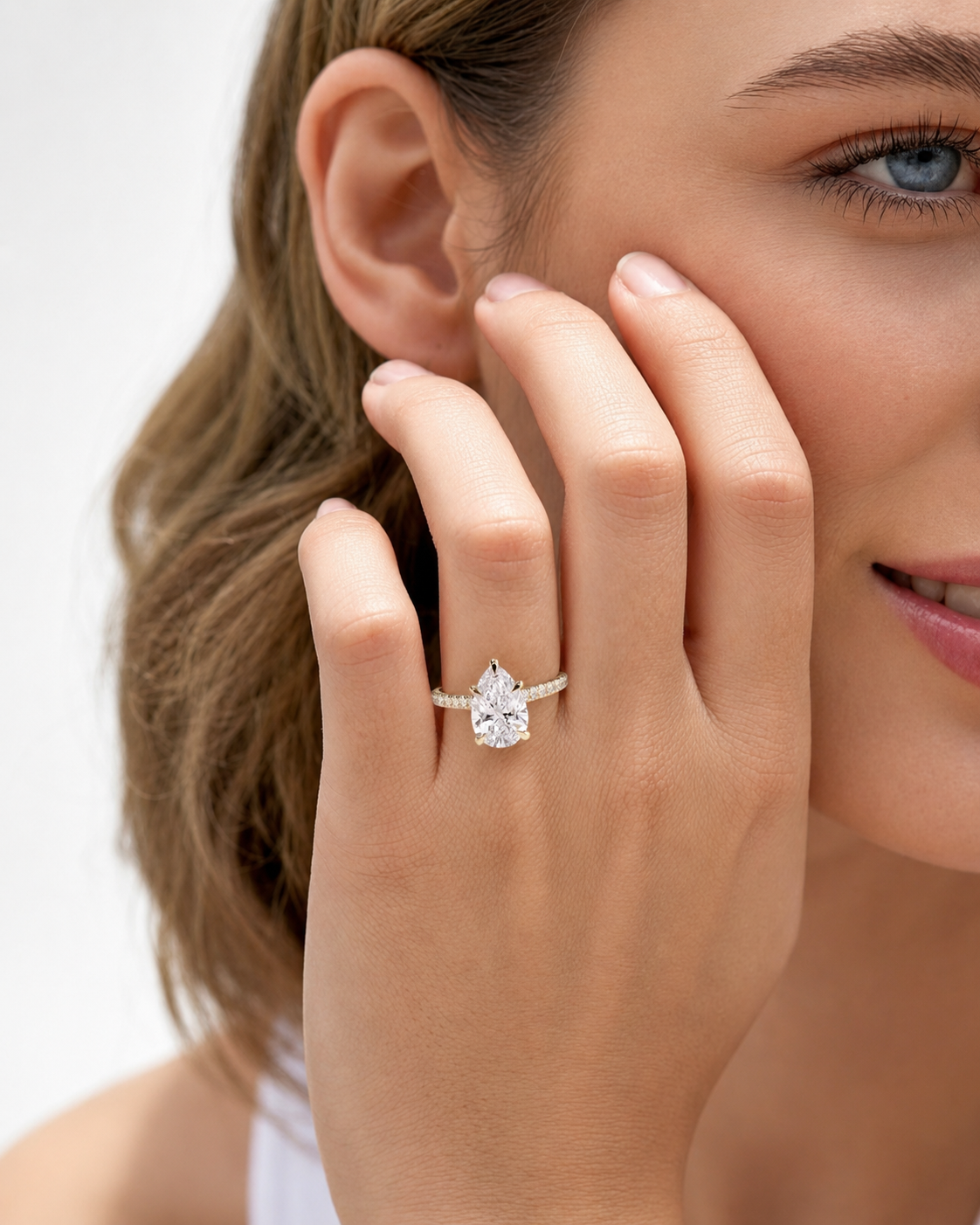 color_10K_YG, color_14K_YG &&A close-up lifestyle shot of a woman wearing a pear-cut diamond engagement ring on a gold band, with her hand resting against her cheek.