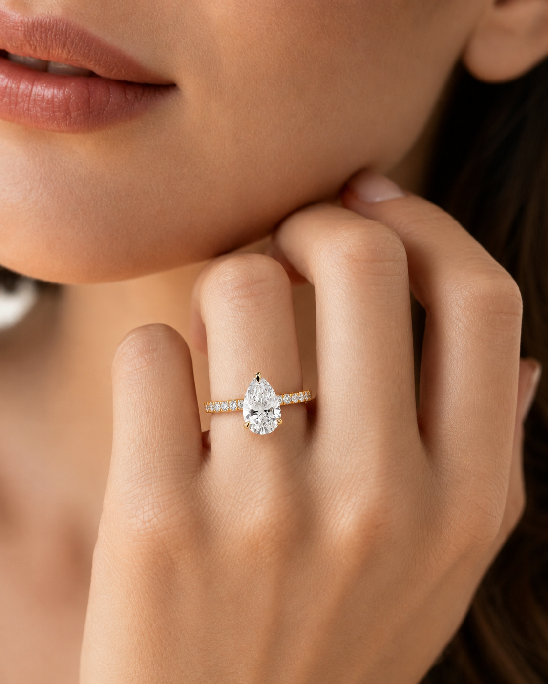 color_10K_YG, color_14K_YG &&A close-up lifestyle shot of a model wearing a pear-cut diamond engagement ring on a gold band with small pavé diamonds.