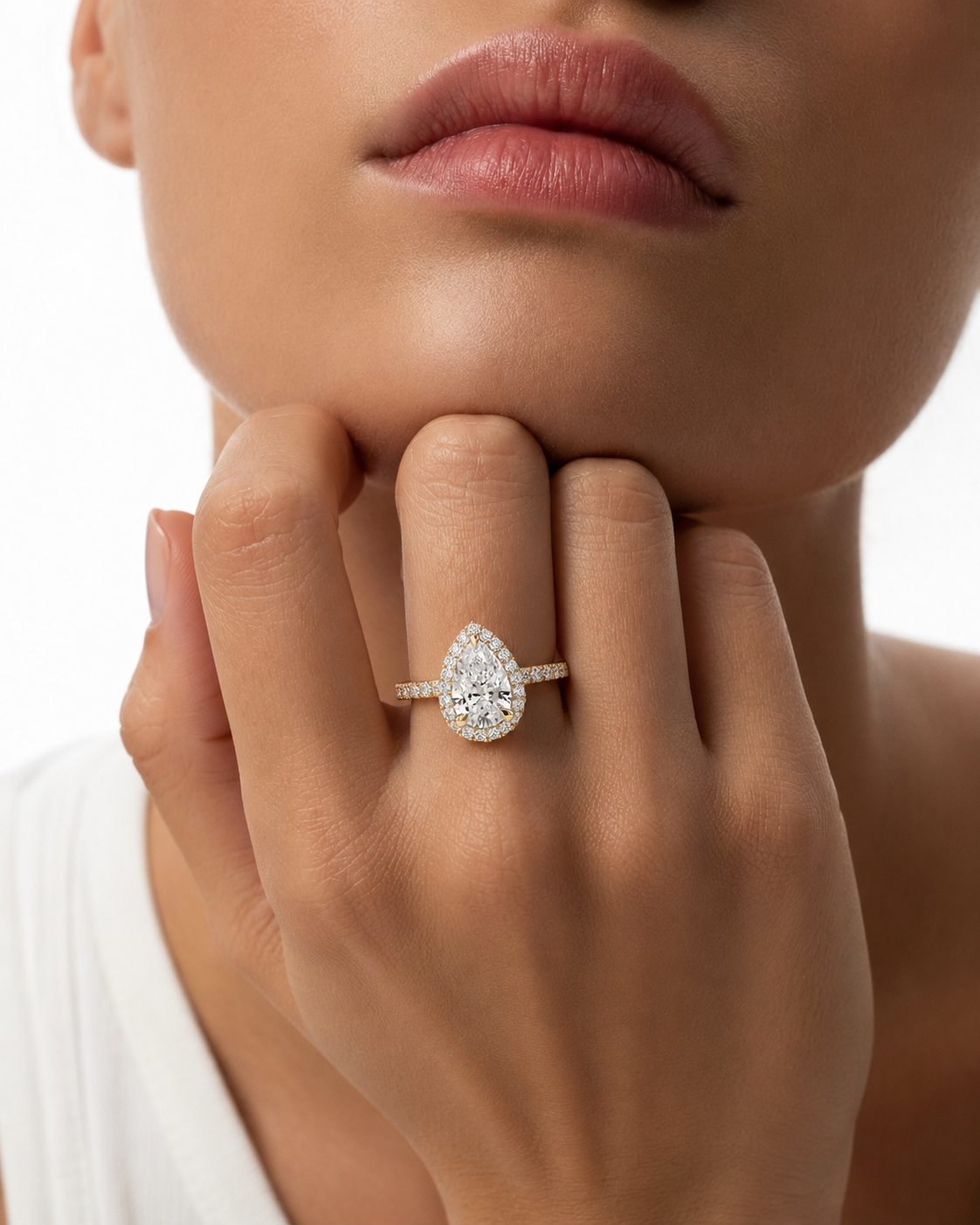 color_10K_YG, color_14K_YG &&A close-up lifestyle shot of a model wearing a pear-cut diamond engagement ring on a gold band, with her hand resting near her chin against a clean white background.