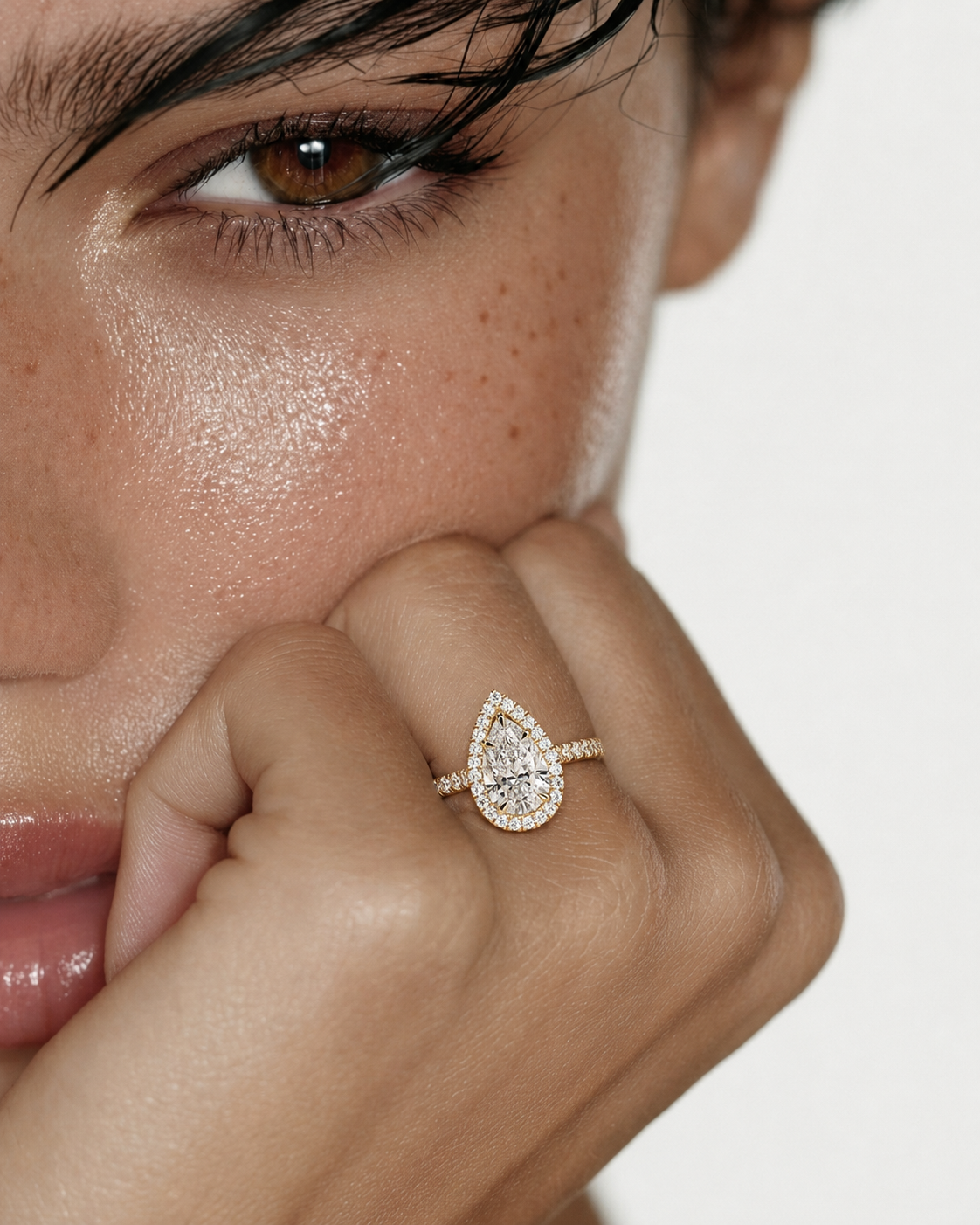color_10K_YG, color_14K_YG &&A close-up lifestyle shot of a model wearing a teardrop-shaped diamond ring with a halo setting on a gold band, resting her chin on her hand against a clean white background.