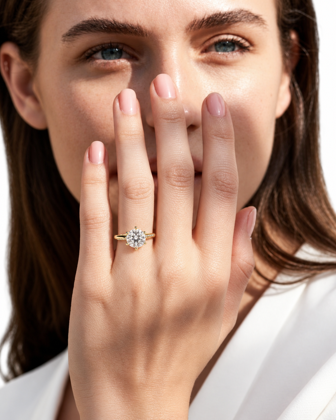 color_10K_YG, color_14K_YG &&A close-up lifestyle shot of a woman wearing a yellow gold halo ring with a clear round-cut center stone on one of her fingers, with her hand covering part of her face.