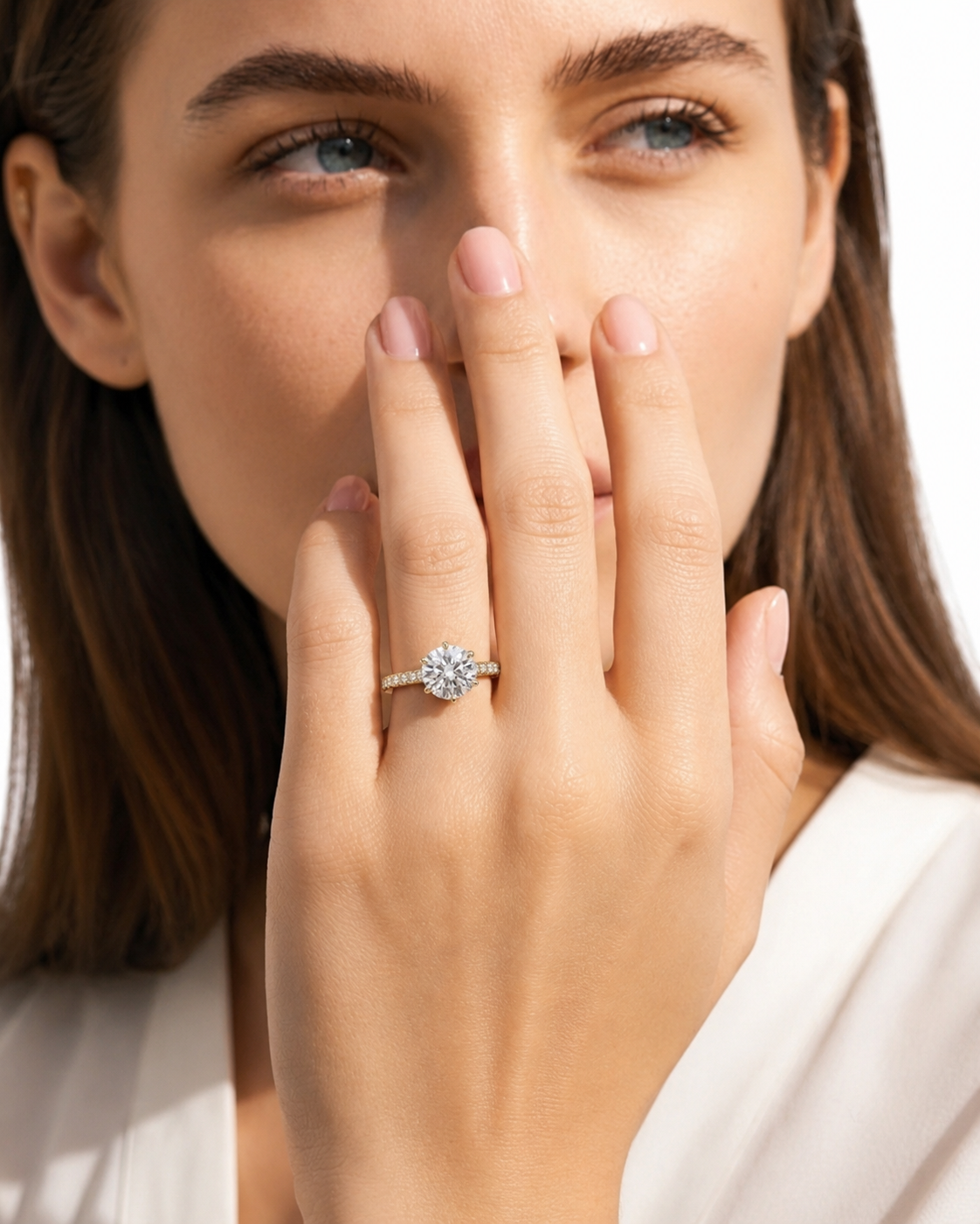 color_10K_YG, color_14K_YG &&A close-up lifestyle shot of a woman wearing a diamond engagement ring on her finger, with her hand covering part of her face against a bright white background.