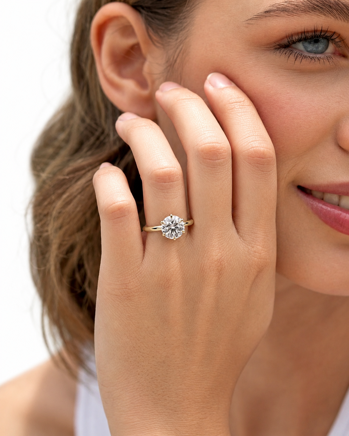 color_10K_YG, color_14K_YG &&A close-up lifestyle shot of a woman wearing a classic solitaire diamond engagement ring on her finger, with her hand resting against her cheek against a clean white background.
