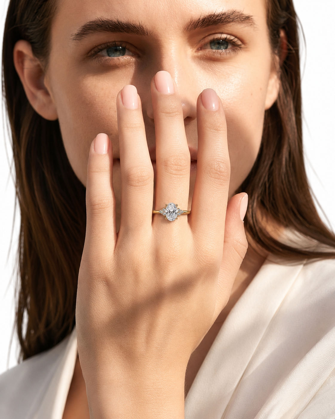 color_10K_YG, color_14K_YG &&A close-up lifestyle shot of a woman wearing a delicate gold ring with a round clear gemstone on her finger, with her hand covering part of her face against a bright white background.