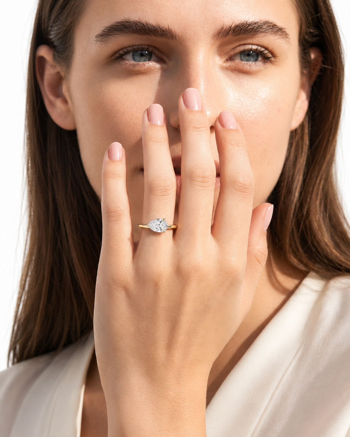 color_10K_YG, color_14K_YG &&A close-up lifestyle shot of a woman holding her hand near her face, showcasing a delicate gold ring with a marquise-cut clear gemstone. The ring is worn on her middle finger, with a second gold band ring on her index finger.