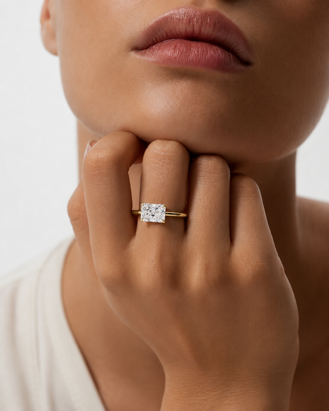 color_10K_YG, color_14K_YG &&A close-up lifestyle shot of a model wearing a yellow gold ring with a square-cut clear gemstone on her finger, with her hand resting on her chin against a clean white background.