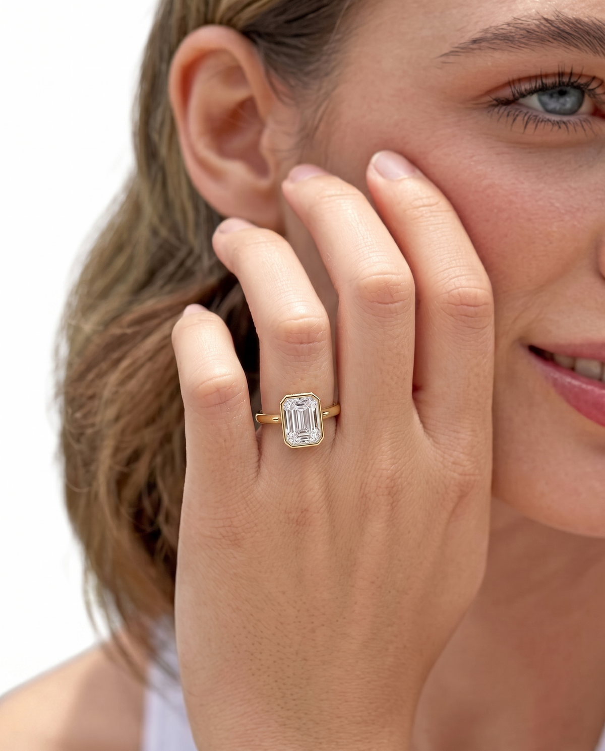 color_10K_YG, color_14K_YG &&
A close-up lifestyle shot of a woman wearing a gold ring with an emerald-cut clear gemstone on her finger, set against a clean white background.