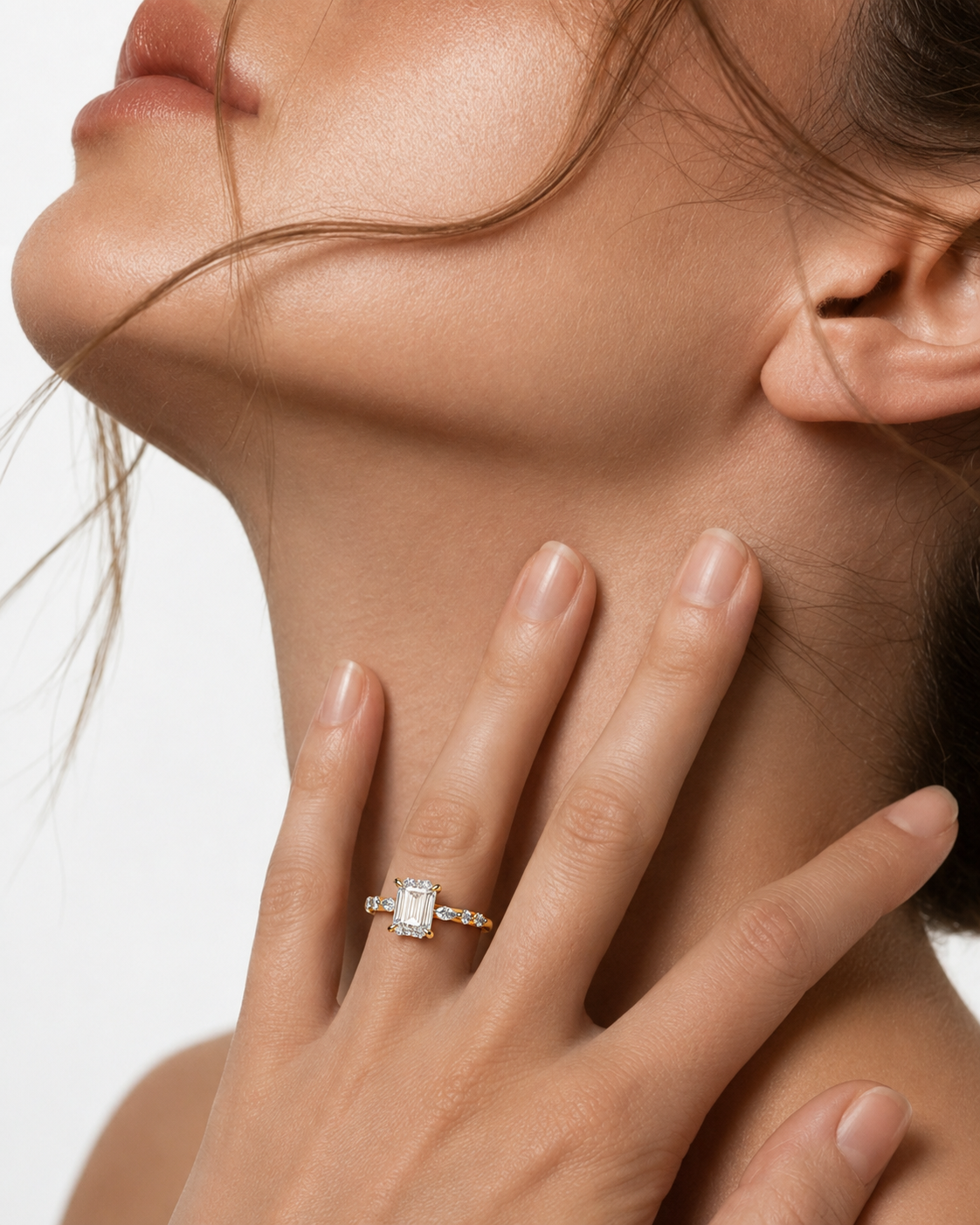 color_10K_YG, color_14K_YG &&A close-up lifestyle shot of a model wearing a yellow gold engagement ring with a rectangular emerald-cut diamond. The model's hand rests gently on her neck against a clean white background.