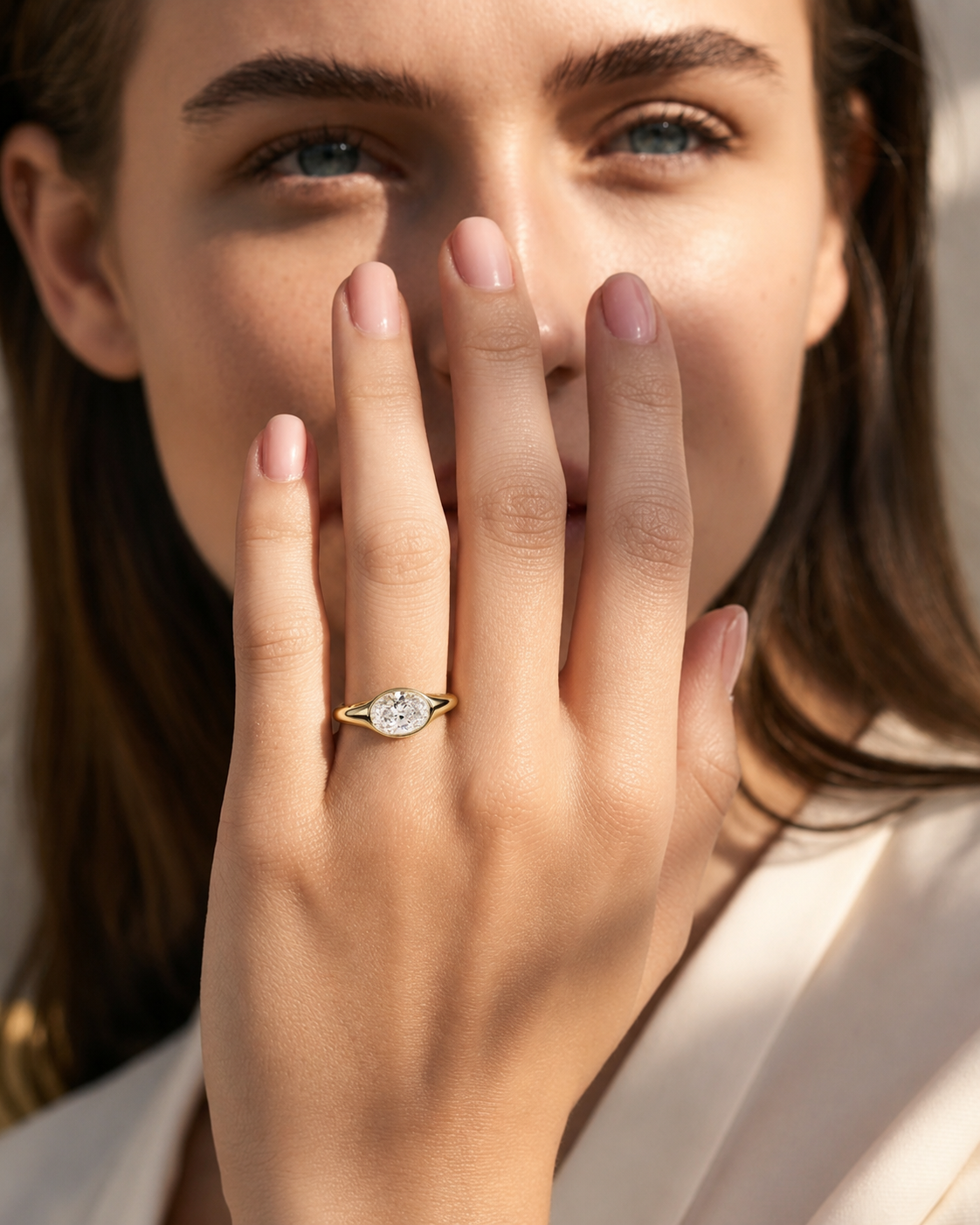 color_10K_YG, color_14K_YG &&A close-up lifestyle shot of a woman wearing a gold ring with a round-cut clear gemstone, holding her hand near her face.