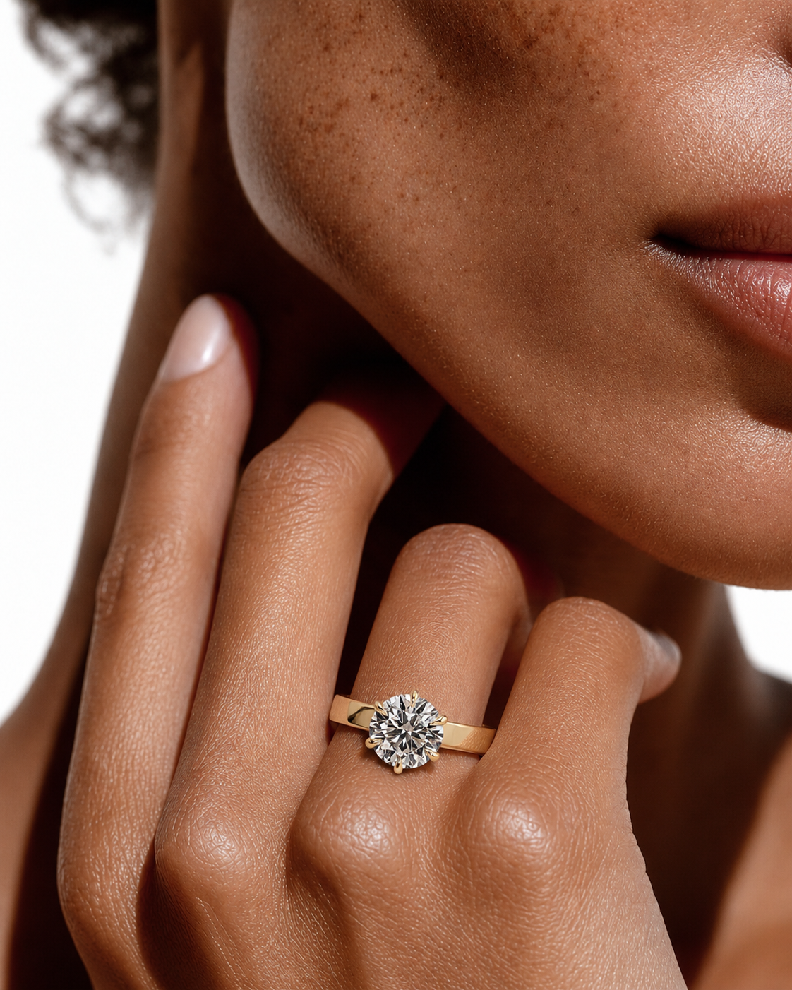 color_10K_YG, color_14K_YG &&A close-up lifestyle shot of a model wearing a yellow gold ring with a round brilliant-cut diamond on her finger. The image features warm, professional lighting and a clean white background.