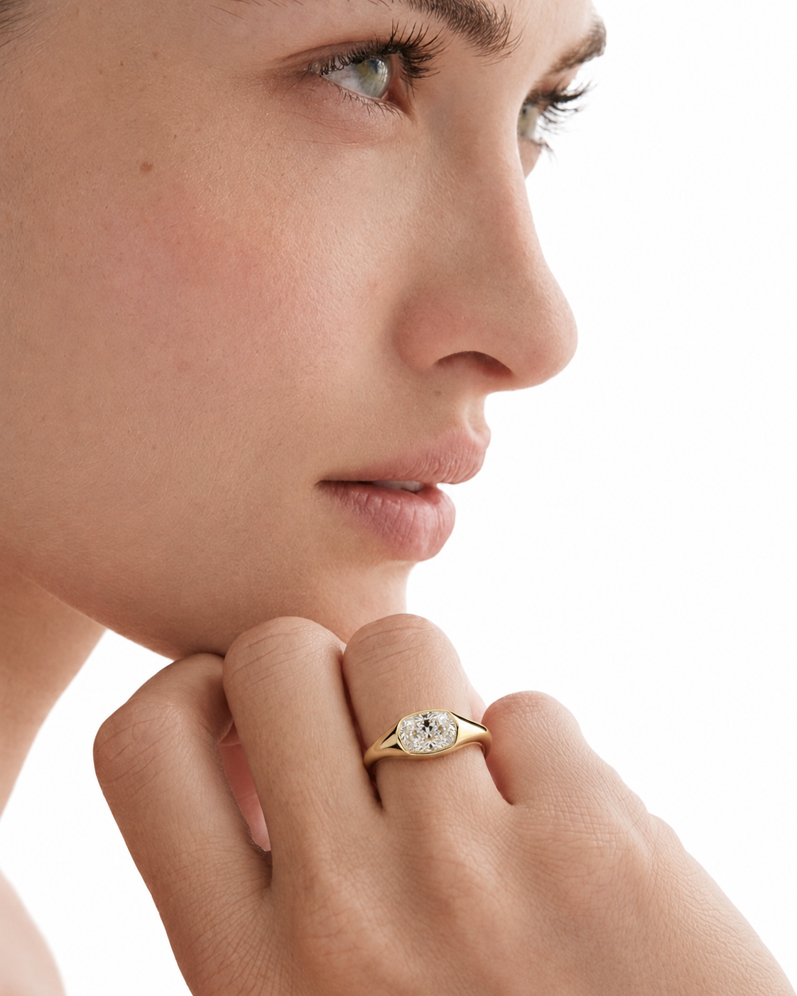 color_10K_YG, color_14K_YG &&A close-up of a model's face and hand wearing a yellow gold ring with a round clear gemstone on a white background.