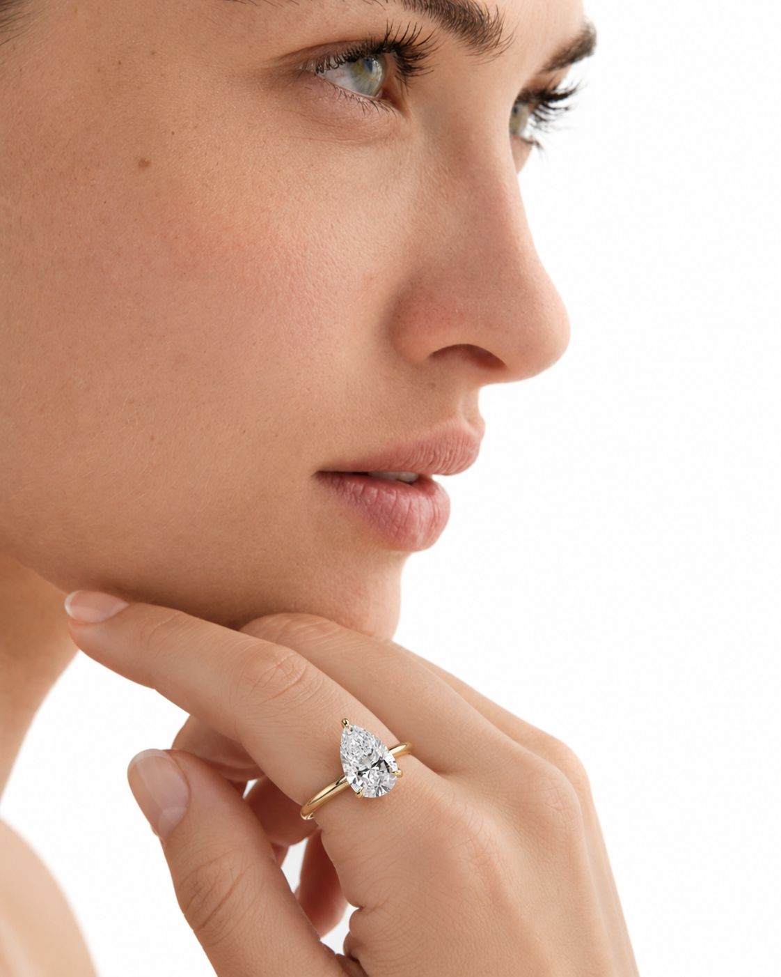 color_10K_YG, color_14K_YG &&A close-up of a woman's face and hand wearing a yellow gold ring with a pear-cut clear gemstone on her ring finger, set against a solid white background.