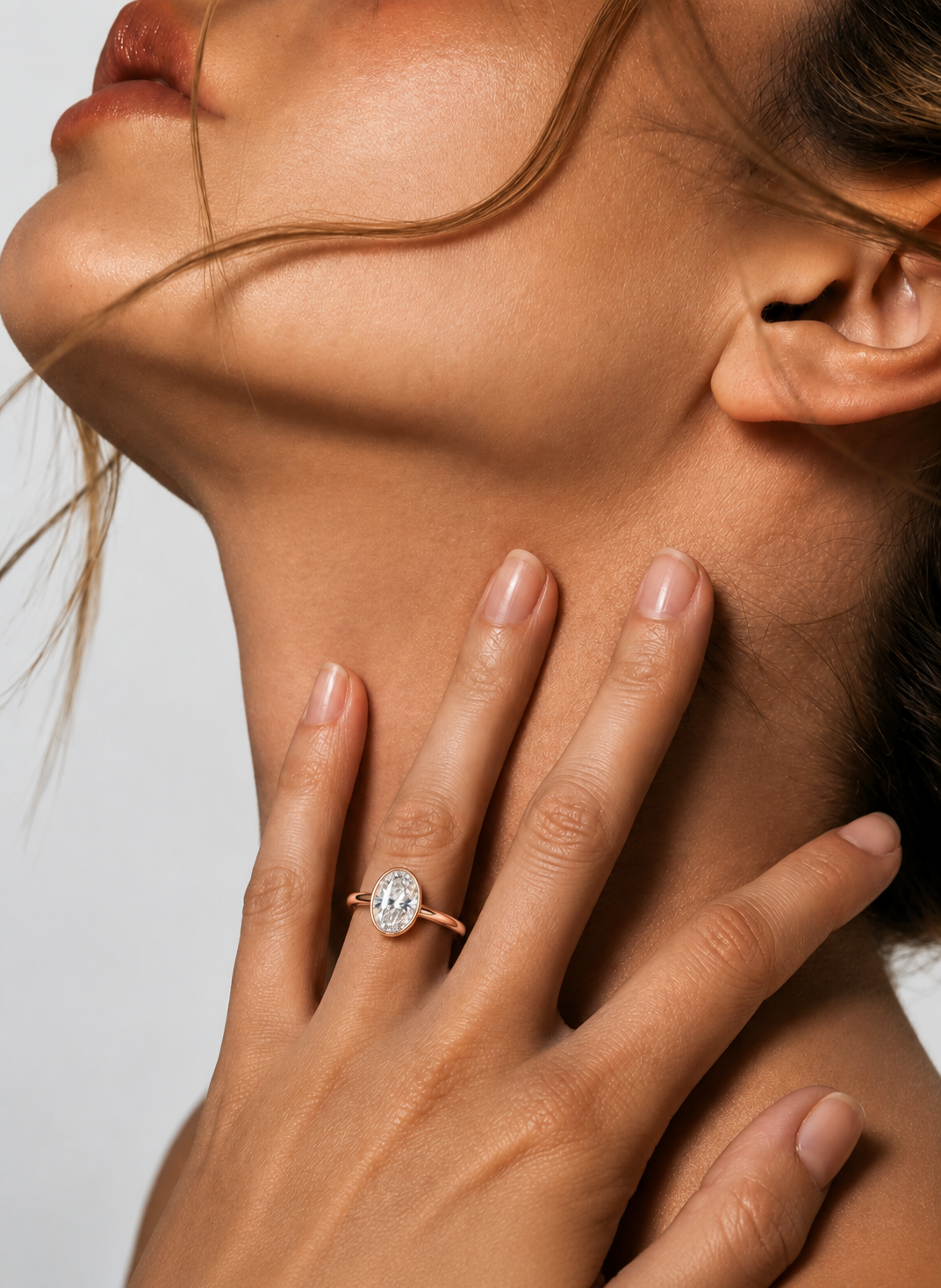 color_10K_RG, color_14K_RG &&
A close-up lifestyle shot of a model wearing a rose gold ring with a clear oval-cut gemstone on her finger, with her hand resting on her neck against a neutral light gray background.