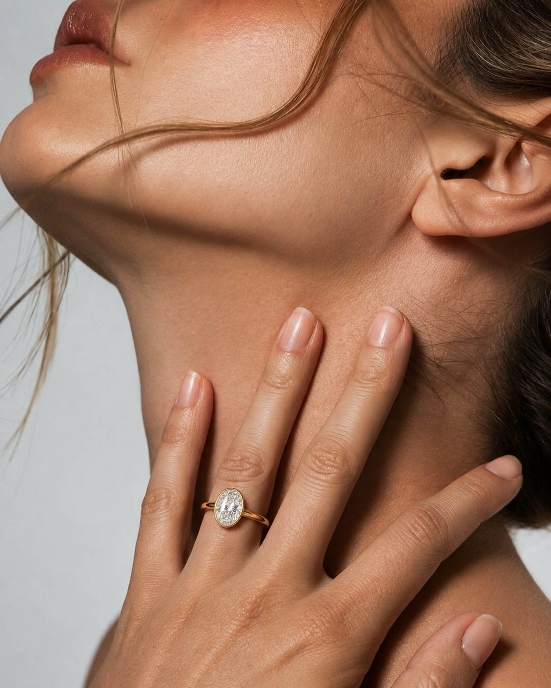 color_10K_YG, color_14K_YG &&
A close-up lifestyle shot of a model wearing a gold ring with a clear oval-cut gemstone on her finger, with her hand resting on her neck against a neutral light gray background.