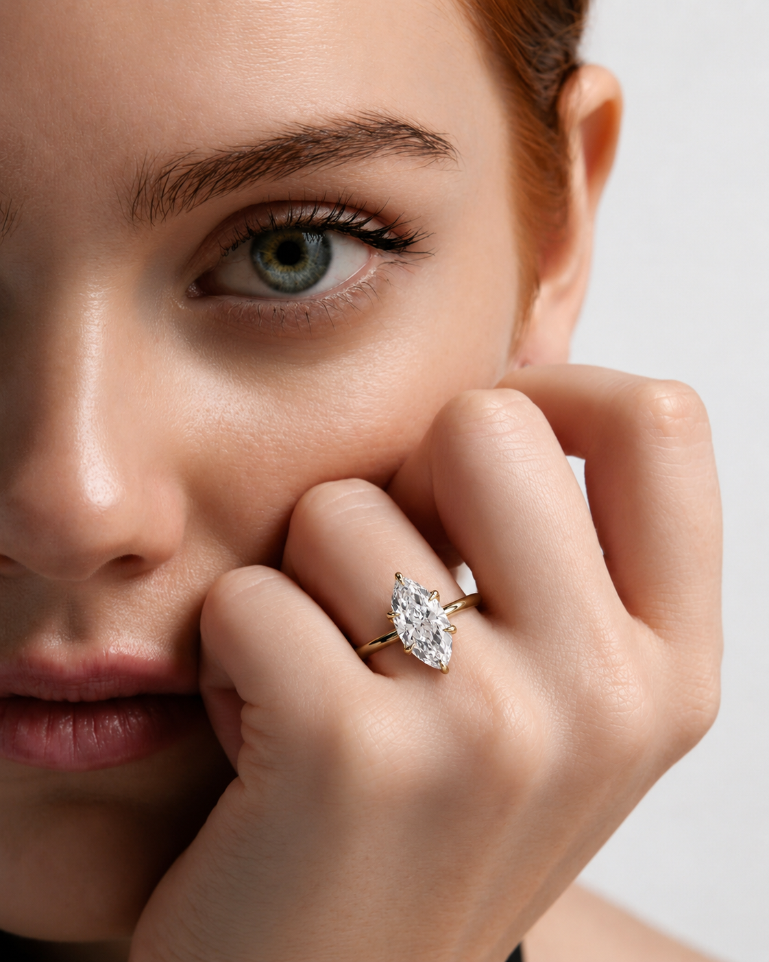 color_10K_YG, color_14K_YG &&A close-up lifestyle shot of a model wearing a gold ring with a marquise-cut clear gemstone on her finger, with her hand resting near her face against a light gray background.