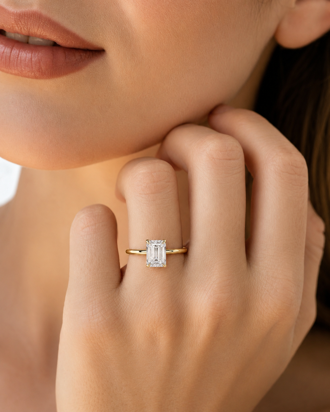 color_10K_YG, color_14K_YG &&A close-up lifestyle shot of a model wearing a yellow gold ring with a rectangular emerald-cut diamond. The ring is shown on her finger, with her hand resting near her chin against a soft, warm background.