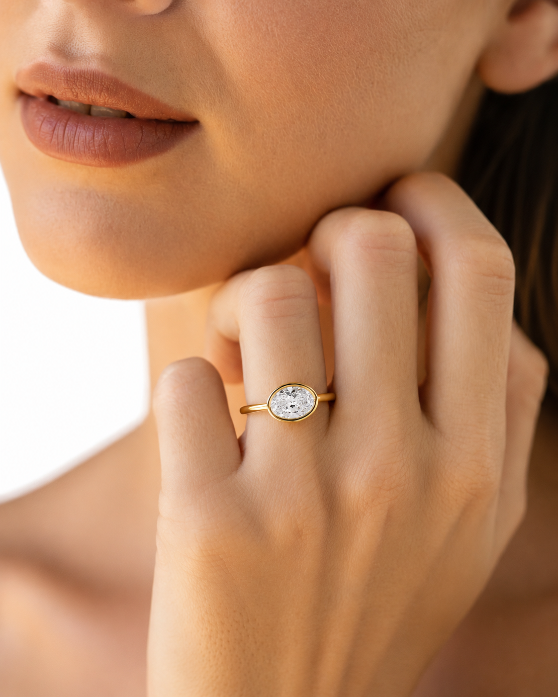 color_10K_YG, color_14K_YG &&A close-up lifestyle shot of a woman's hand wearing a delicate gold ring with a round clear gemstone, set against her face and hand.