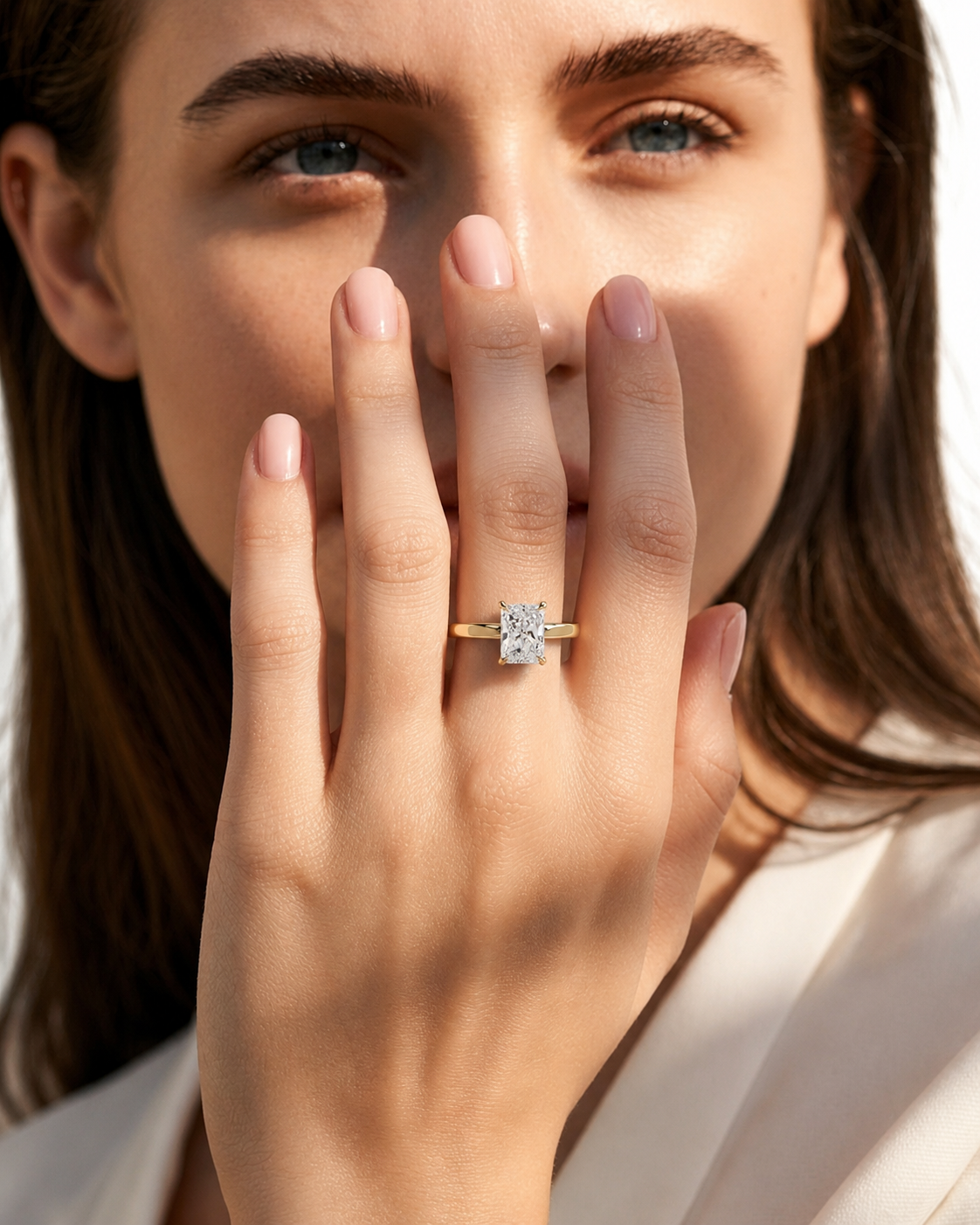 color_10K_YG, color_14K_YG &&A close-up lifestyle shot of a woman wearing a yellow gold ring with a square-cut clear gemstone on her finger, with her hand covering part of her face.