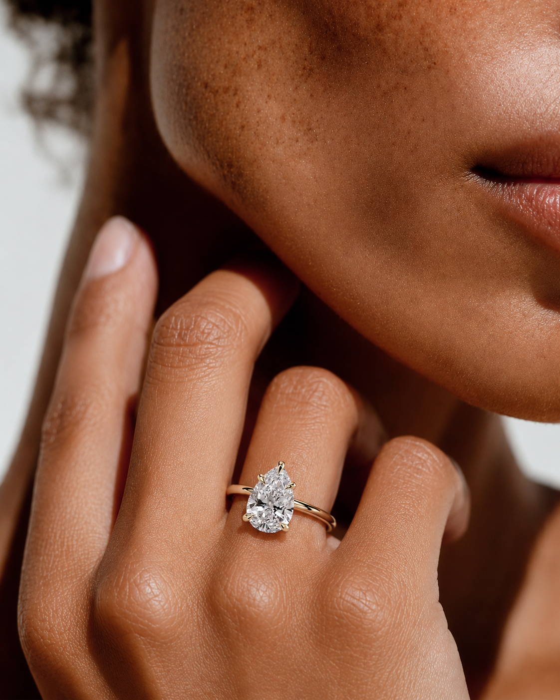 color_10K_YG, color_14K_YG &&
A close-up lifestyle shot of a model wearing a pear-cut diamond ring on a gold band, with her hand resting near her face.
