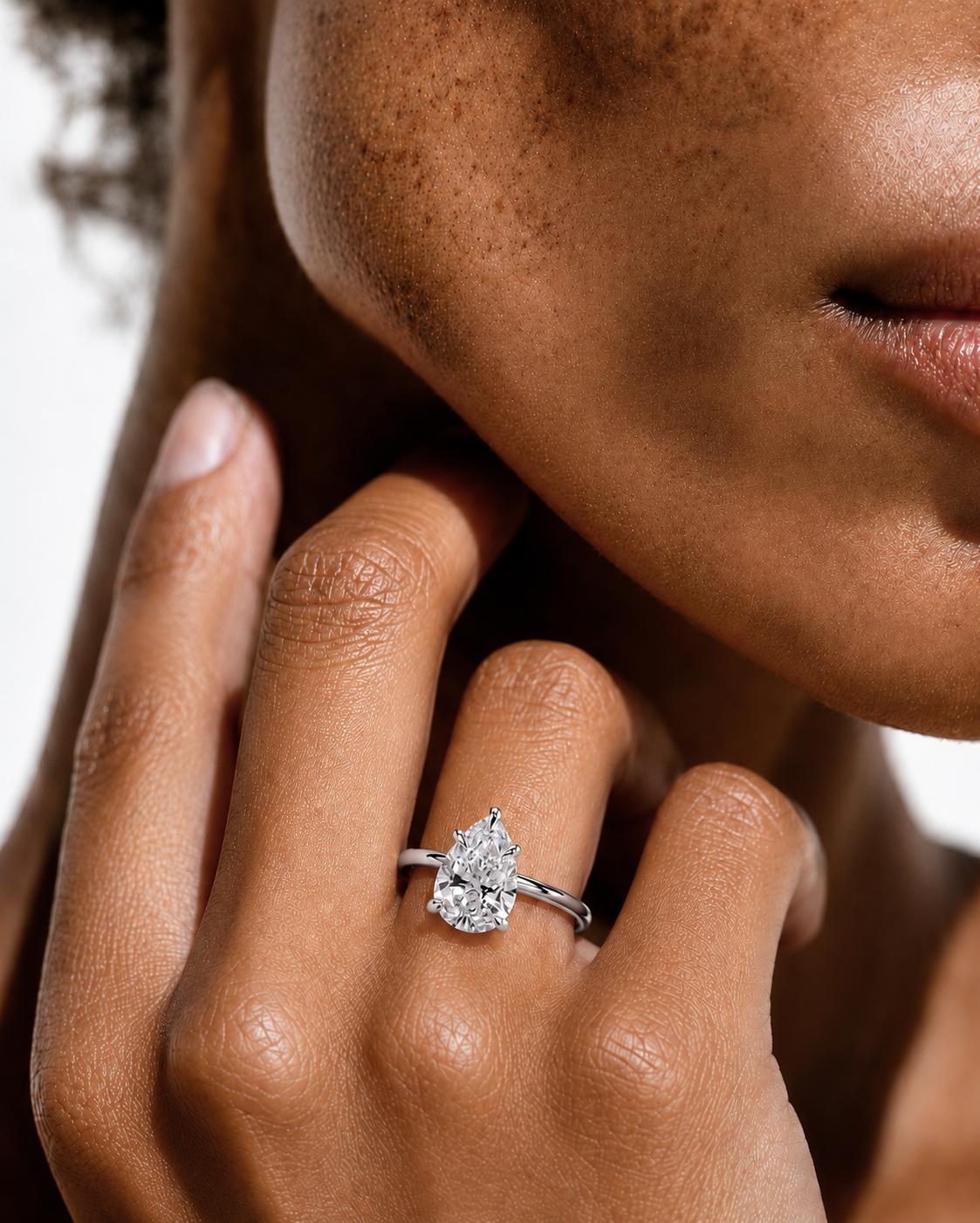 color_925_SIL,color_935_ARG,color_10K_WG, color_14K_WG &&
A close-up lifestyle shot of a model wearing a silver band ring with a pear-cut clear gemstone on her finger, with her hand resting near her face.
