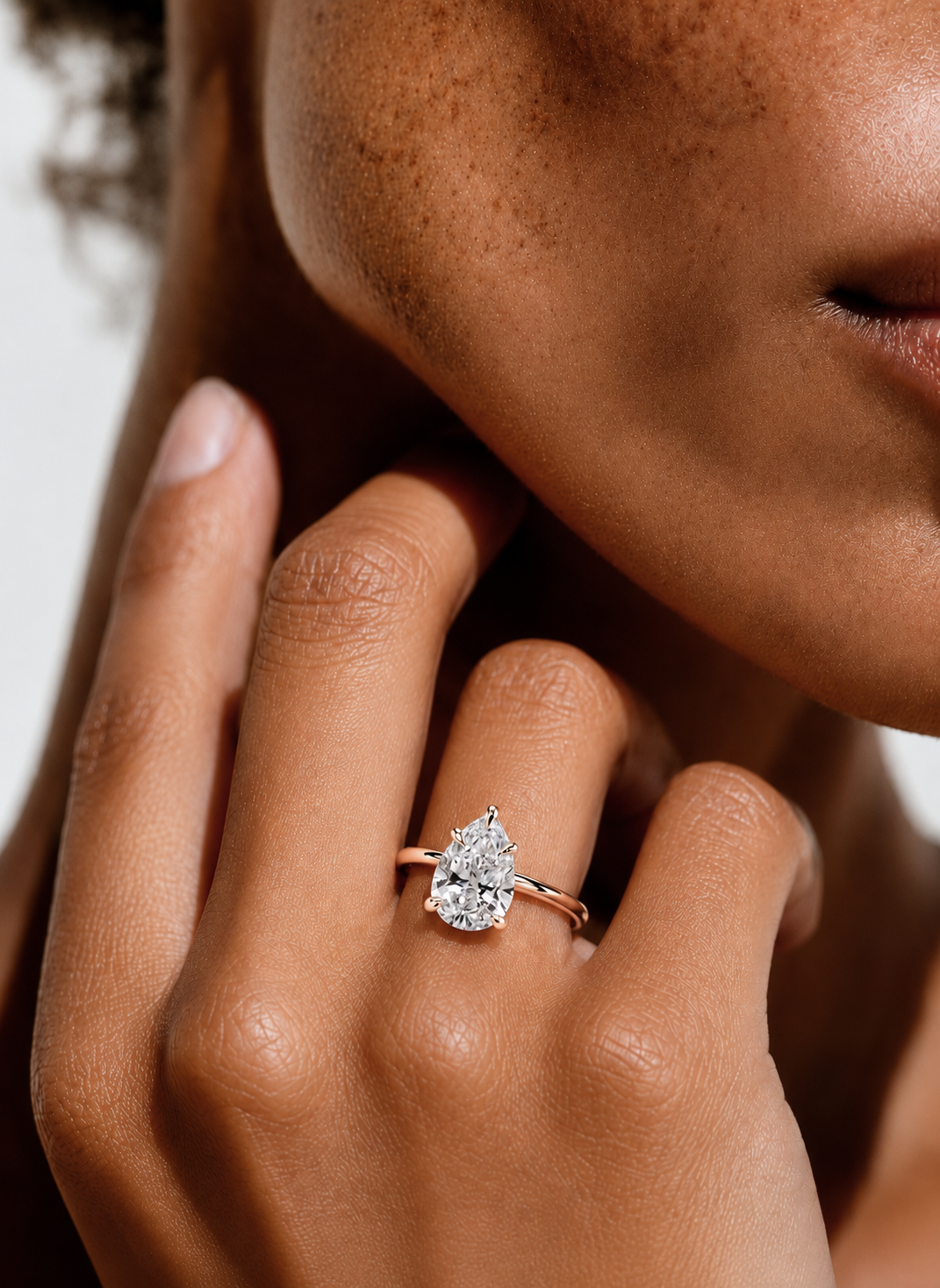 color_10K_RG, color_14K_RG &&
A close-up lifestyle shot of a model wearing a pear-cut diamond engagement ring on a rose gold band.