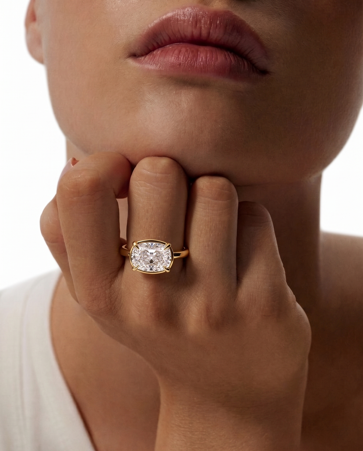 color_10K_YG, color_14K_YG &&A close-up lifestyle shot of a model wearing a yellow gold ring with a cushion-cut diamond on her finger, with her hand resting on her chin against a white background.