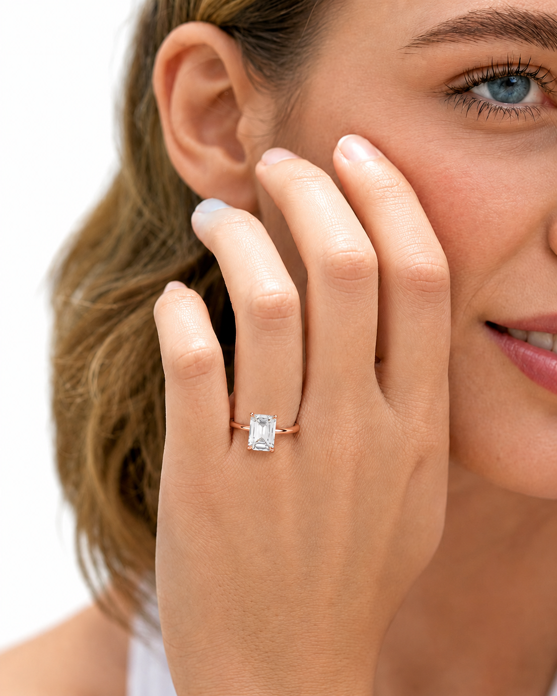 color_10K_RG, color_14K_RG &&
A close-up lifestyle shot of a woman wearing a rose gold ring with a rectangular clear gemstone on her finger, with her hand resting against her cheek against a white background.