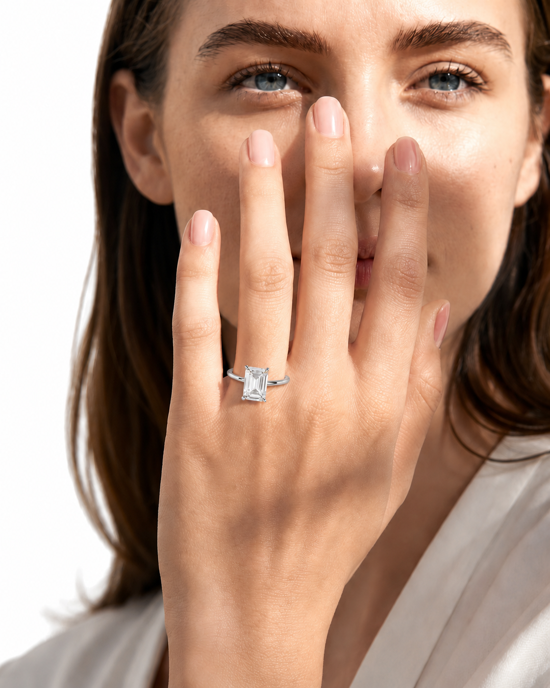 color_925_SIL,color_935_ARG,color_10K_WG, color_14K_WG &&
A close-up lifestyle shot of a woman wearing a silver engagement ring with an emerald-cut clear center stone on her ring finger, with her hand covering part of her face against a white background.