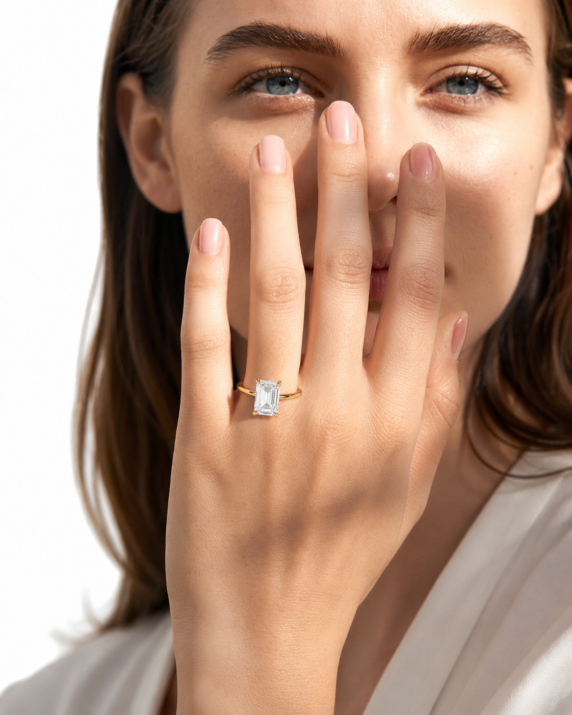 color_10K_YG, color_14K_YG &&
A close-up lifestyle shot of a woman wearing a yellow gold ring with an emerald-cut clear gemstone on her finger, holding her hand near her face against a bright white background.