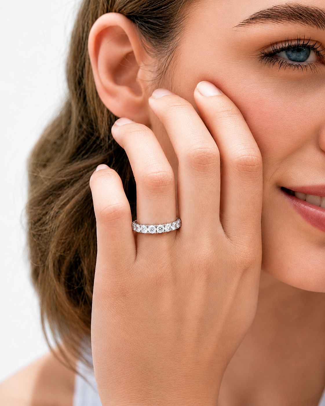 color_925_SIL,color_935_ARG,color_10K_WG, color_14K_WG &&	
A close-up lifestyle shot of a woman wearing a silver eternity band ring with clear round-cut gemstones on her finger, with her hand resting against her cheek.