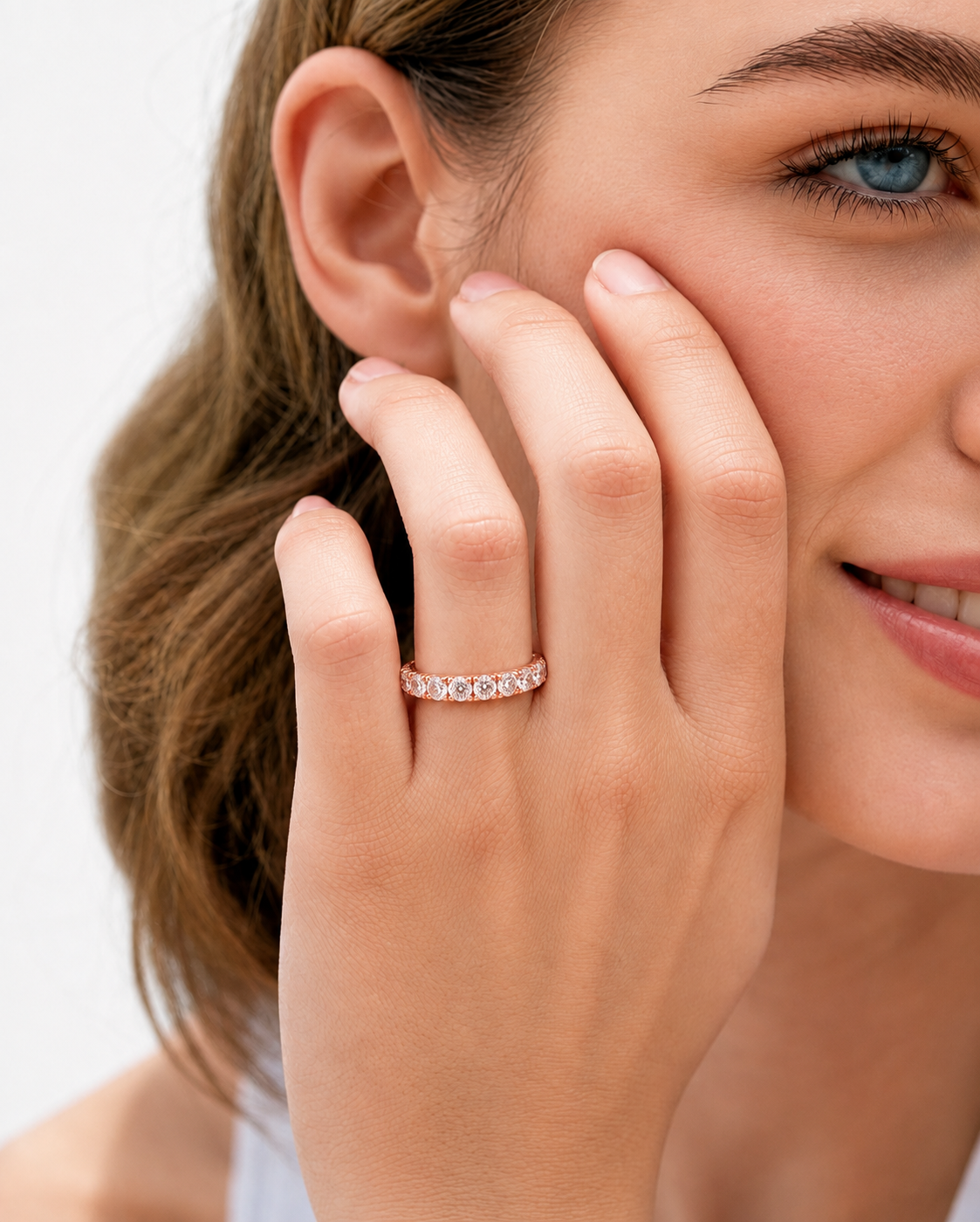 color_10K_RG, color_14K_RG &&	A close-up lifestyle shot of a woman wearing a delicate rose gold eternity ring with small round-cut stones on her ring finger, with her hand resting against her cheek.