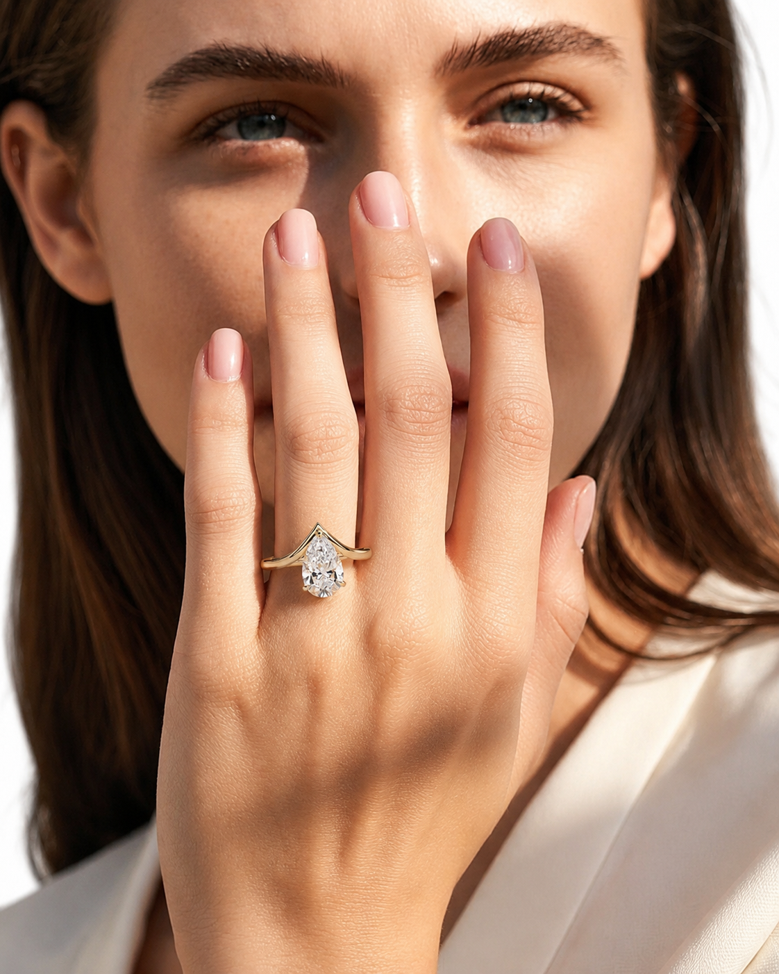 color_10K_YG, color_14K_YG &&A close-up lifestyle shot of a woman wearing a yellow gold ring with a pear-cut clear gemstone on her finger, holding her hand near her face against a bright white background.
