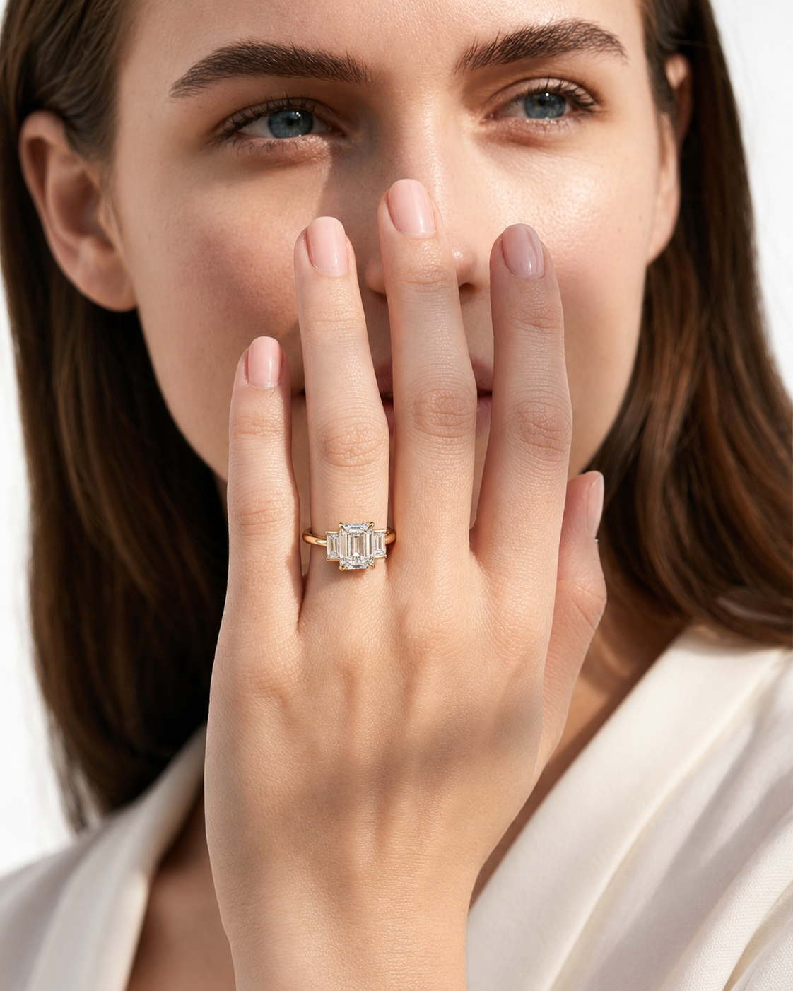 color_10K_YG, color_14K_YG &&A close-up lifestyle shot of a woman wearing a gold ring with a large emerald-cut clear center stone and smaller side stones on her finger, with her hand covering part of her face.