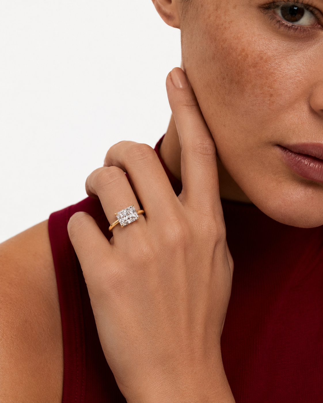 color_10K_YG, color_14K_YG &&A close-up lifestyle shot of a model wearing a yellow gold ring with a square-cut clear gemstone. The model is wearing a deep burgundy top, and the background is a clean, solid white.