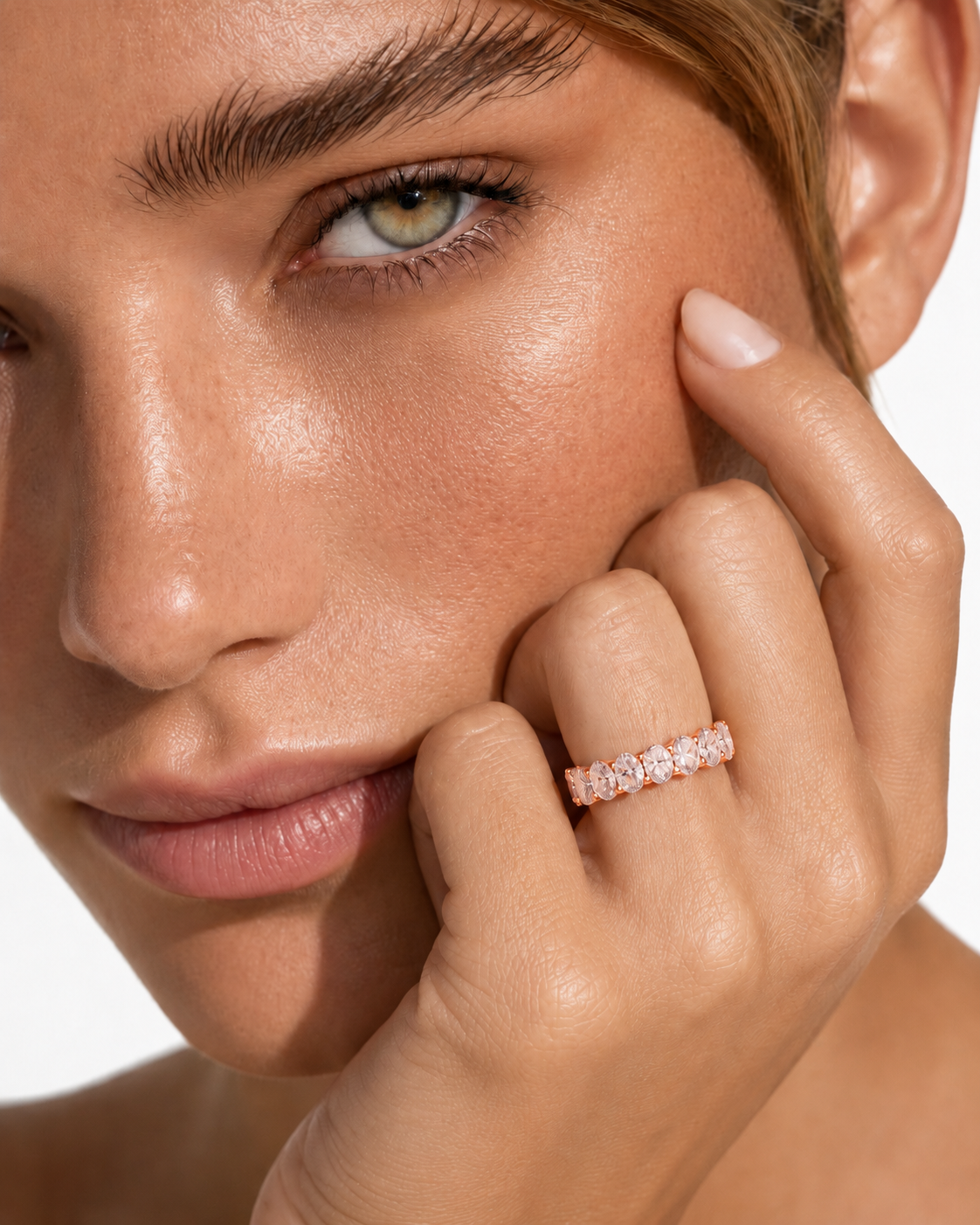 color_10K_RG, color_14K_RG &&	A close-up lifestyle shot of a model wearing a delicate rose gold eternity ring set with small clear stones on her finger, with her hand resting against her cheek against a clean white background.