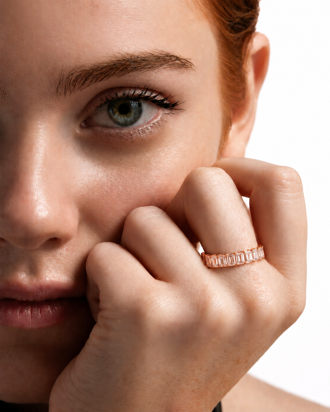 color_10K_RG, color_14K_RG &&	Close-up of a woman resting her chin on her hand, wearing a rose gold band ring with a row of small baguette-cut clear stones.