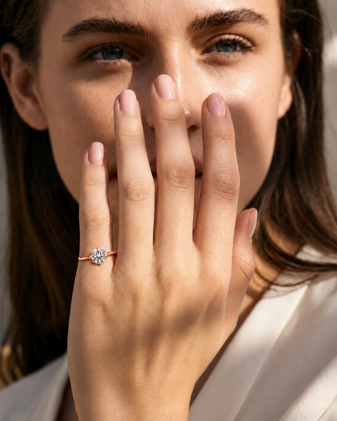 color_10K_RG, color_14K_RG &&A close-up lifestyle shot of a woman holding her hand near her face, showcasing a delicate rose gold ring with a small clear gemstone cluster.